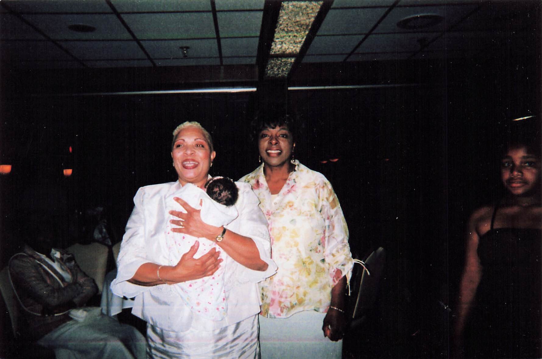 Smiling woman in white holds a newborn baby, standing with another woman at a dimly lit party.