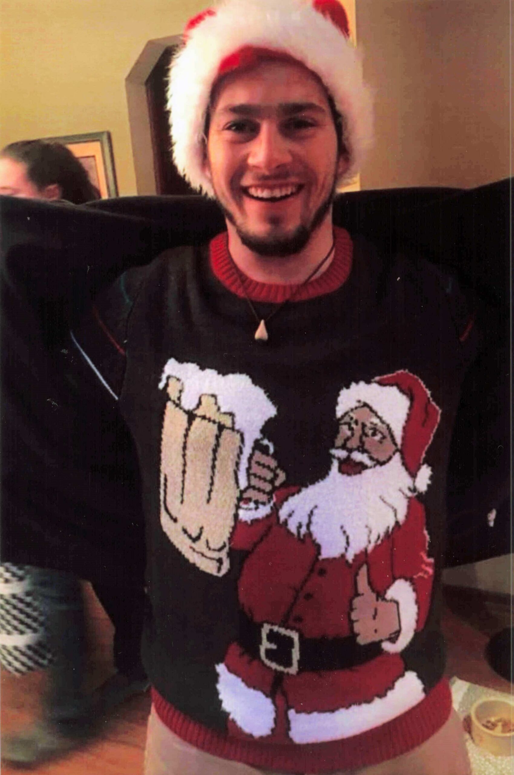 Smiling man wearing a Santa hat and a Christmas sweater with Santa Claus imagery, indoors.