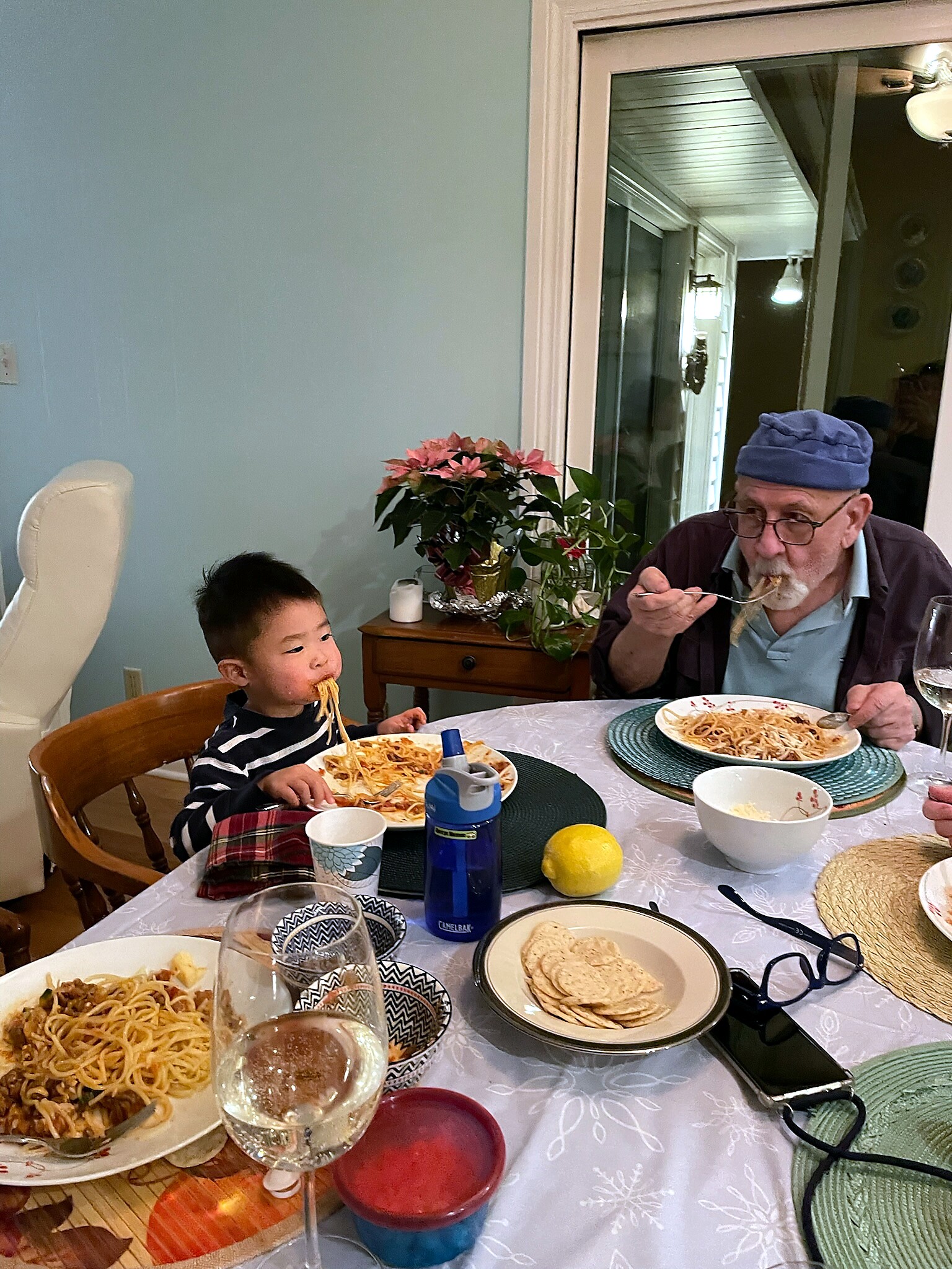 Child and an elderly man dining at a table with spaghetti plates and a lemon on a light-colored tablecloth.