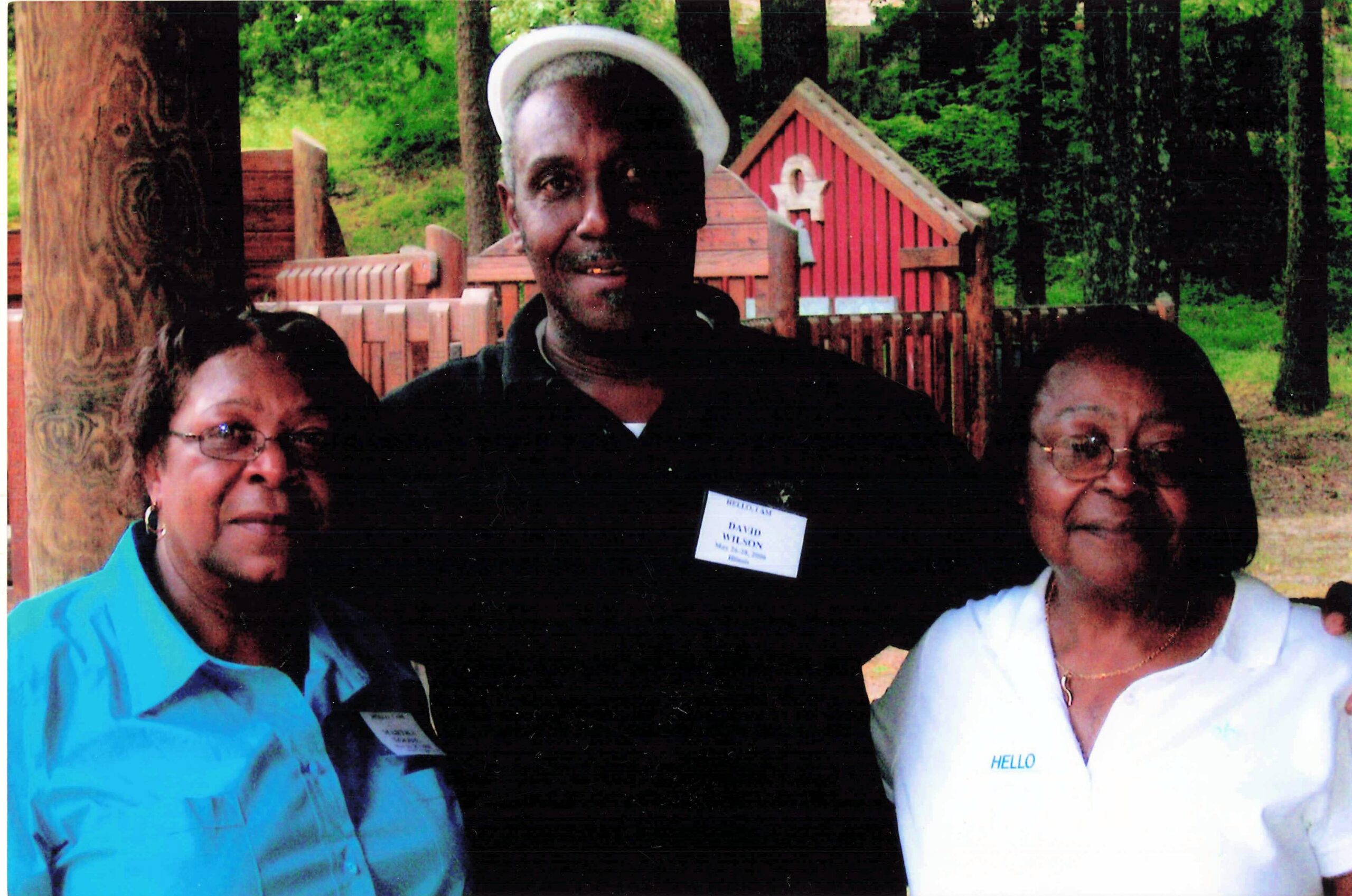 Three adults smile for a photo outdoors, with a wooden fence and a small red playhouse in the background; woman in blue on the left, man in a black shirt and white hat center, woman in white on the right.