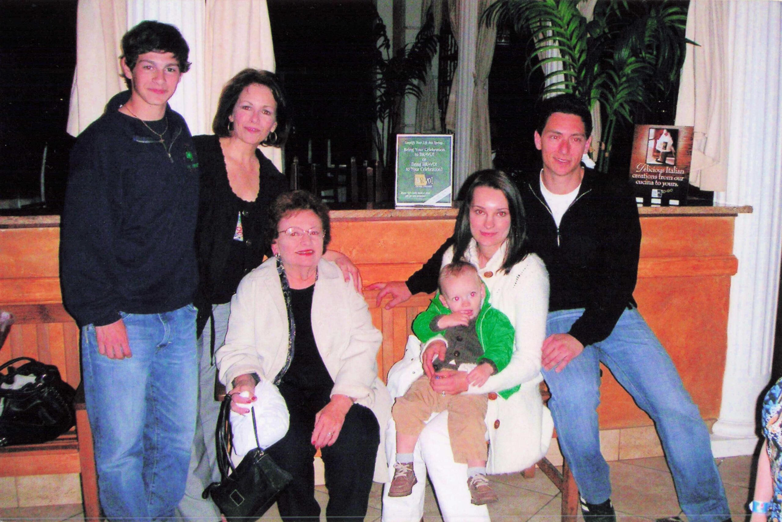 Group of five adults and a baby posing for a photo in a restaurant, gathered around a wooden counter with a baby in a green jacket in front.