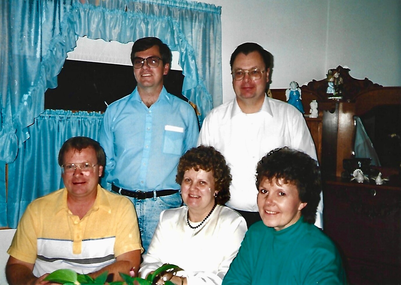 Five adults posing for a casual group photo in a living room with blue curtains behind them.