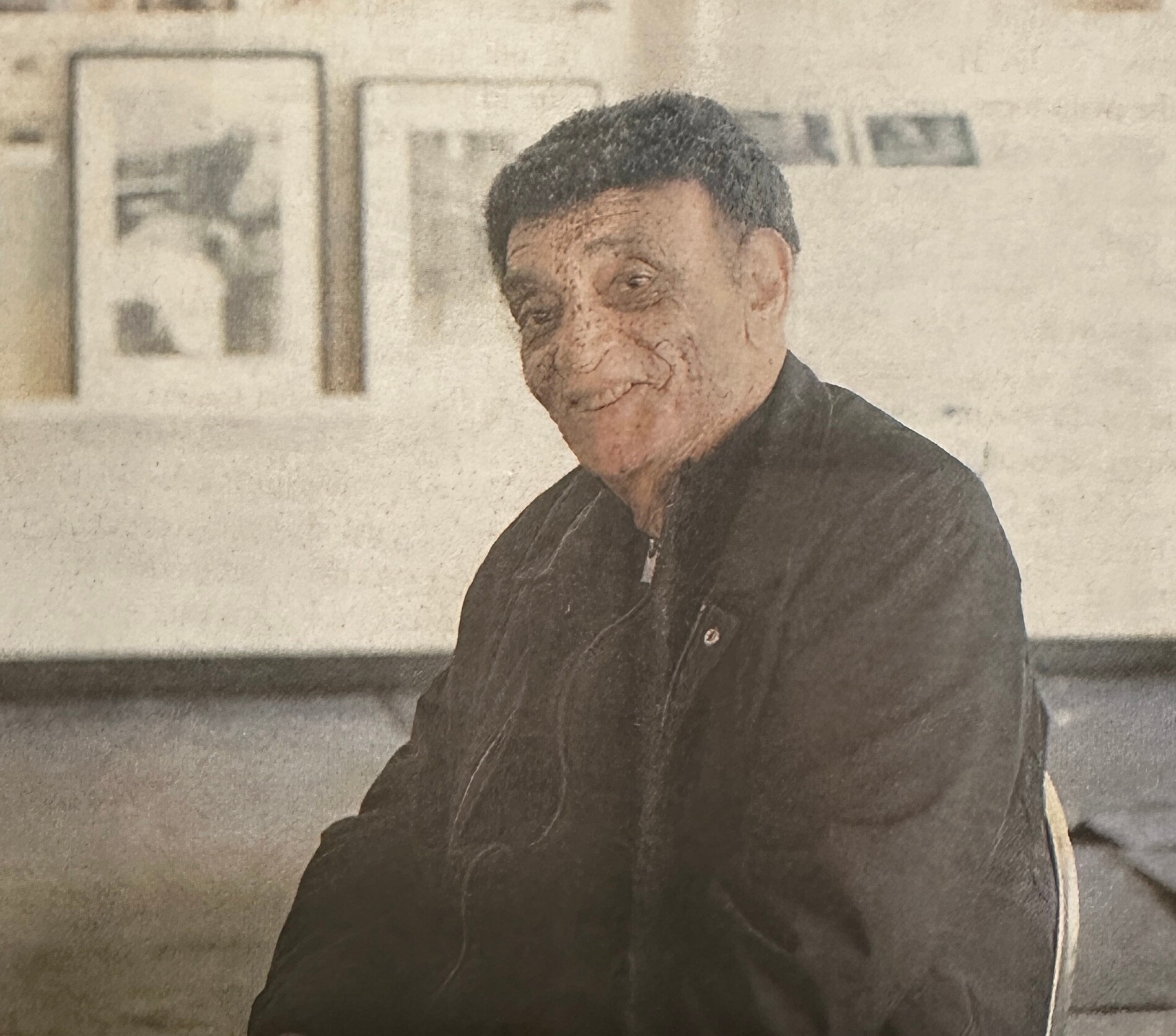 Older man in a dark shirt stands indoors near framed artwork on the wall in a gallery-like setting.