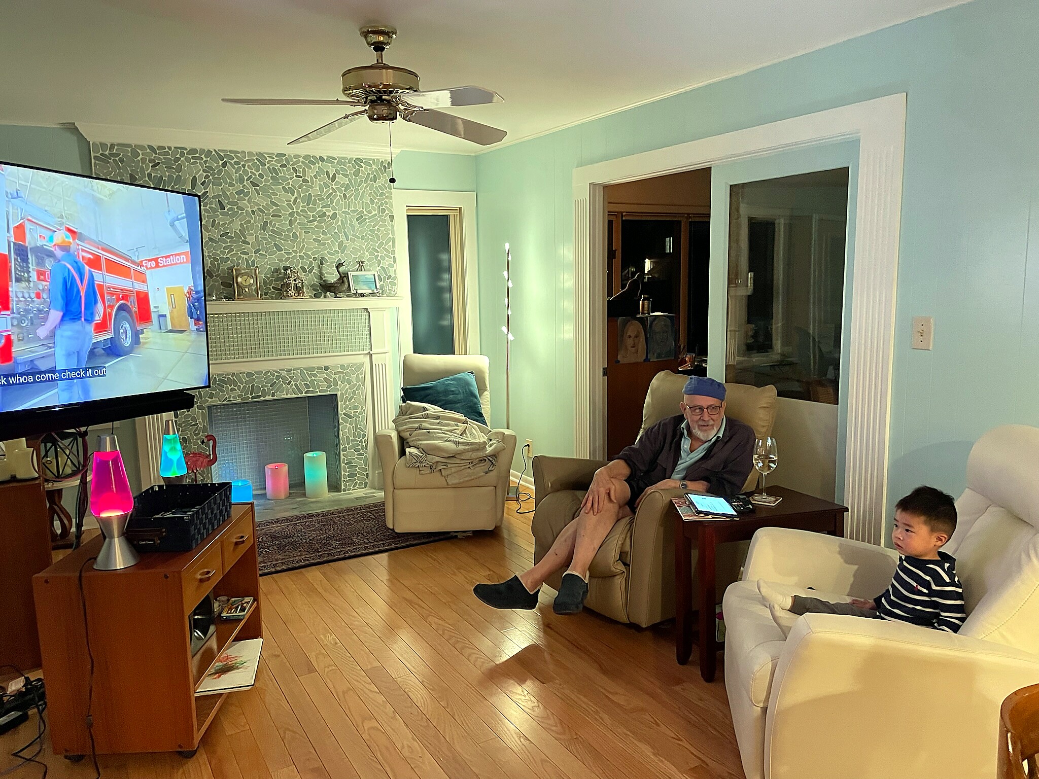 Living room scene with a man in a beige chair watching TV, and a child on a white chair nearby.