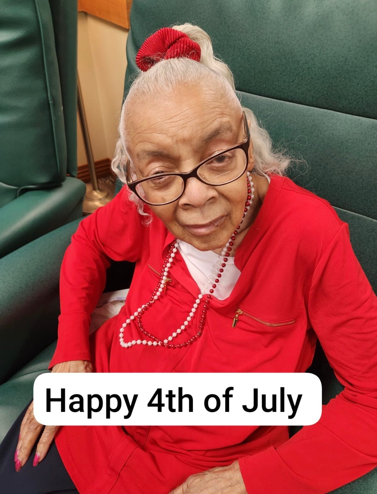 Smiling elderly woman with white hair in a red outfit and beaded necklace, wearing glasses and a red scrunchie, celebrating Independence Day.