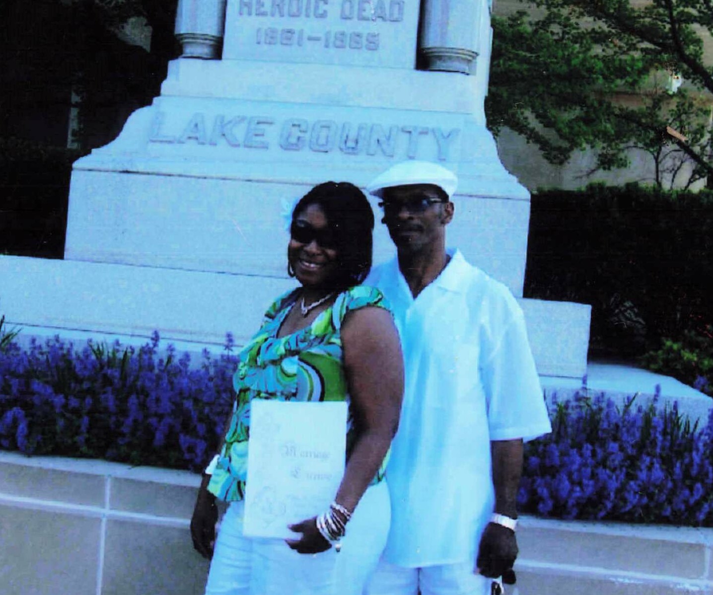 Smiling couple posing in front of a pale Lake County monument, woman in a green print top and white pants, holding a folder.