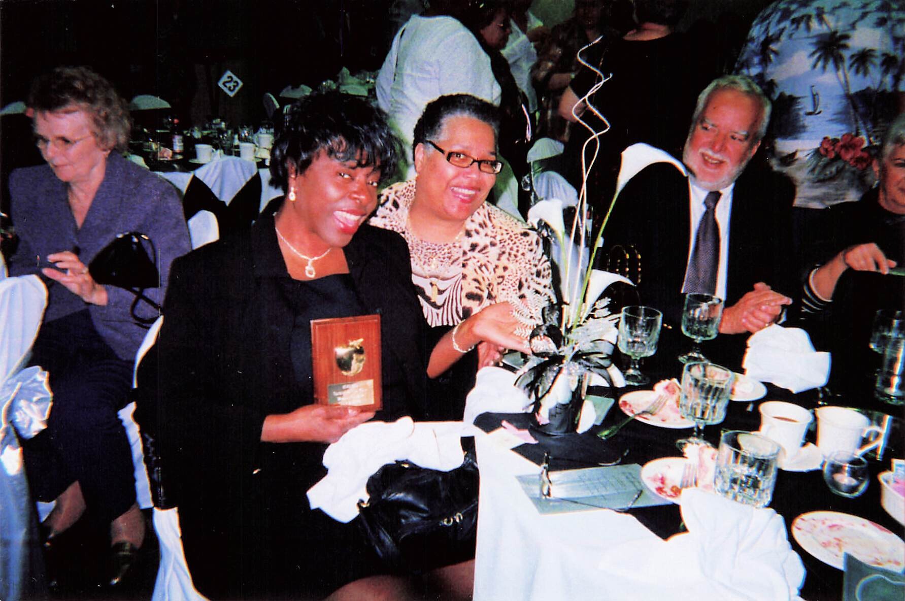 Smiling group at a banquet table; woman center holds a wooden award plaque, centerpiece flowers in front, guests around.