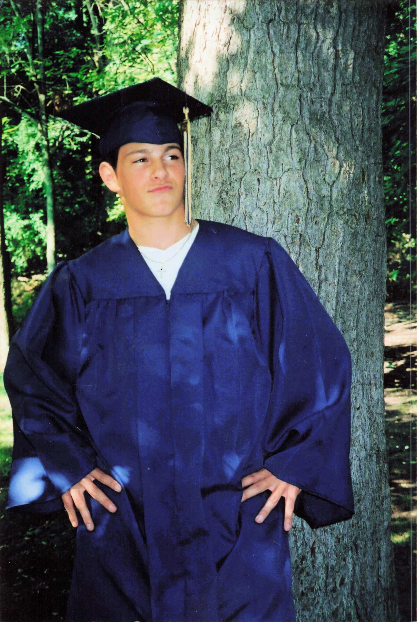Young man in a blue graduation gown and cap poses beside a tree outdoors, after graduation.