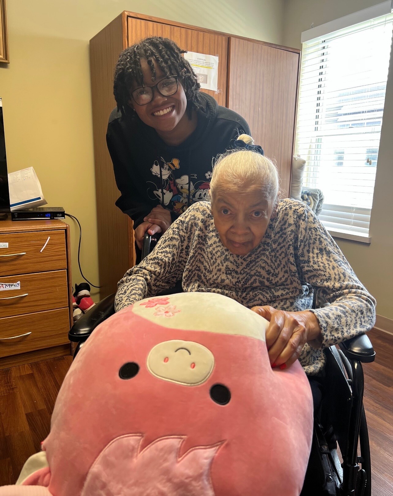 Smiling young caregiver with glasses leans over an elderly woman in a wheelchair inside a sunny room, with a large pink plush toy in the foreground.
