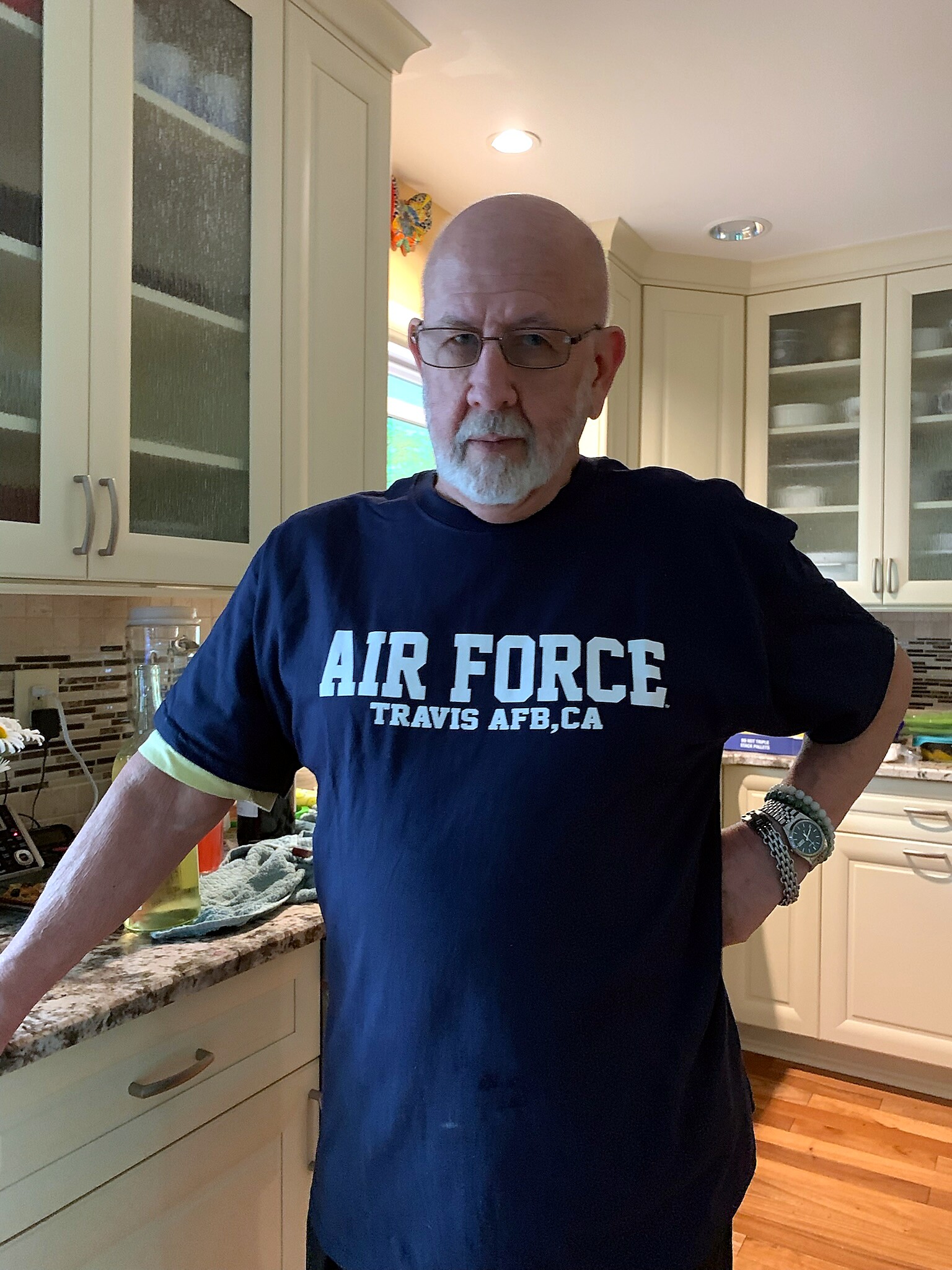 Older man with glasses and a white beard standing in a kitchen wearing a navy Air Force T-shirt that reads 'Travis AFB, CA'.