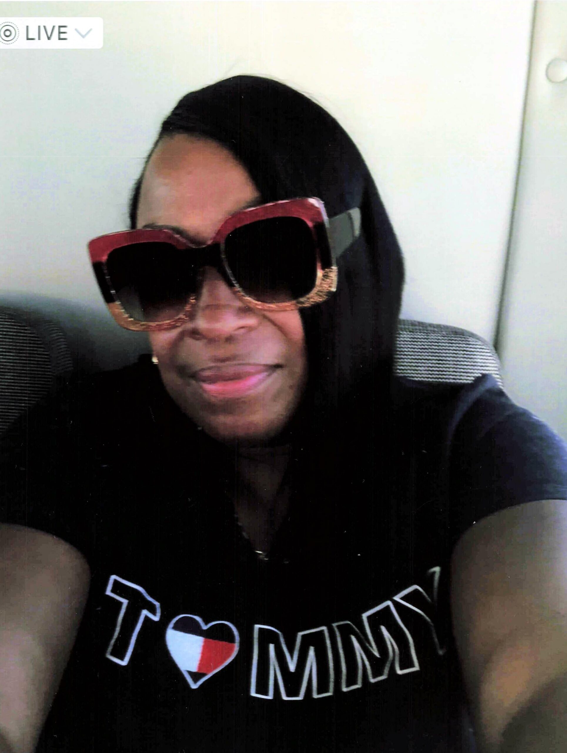 Selfie of a woman wearing a black hijab and oversized red sunglasses, smiling; her shirt shows a graphic text with a heart symbol (I ❤) and larger letters.