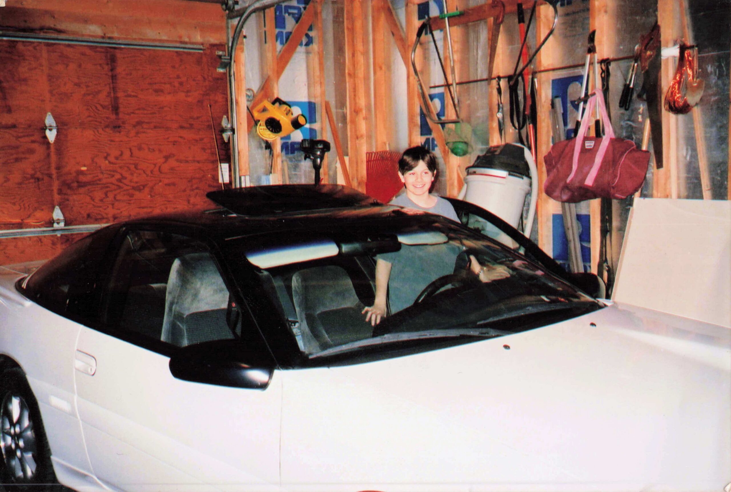 Smiling boy stands beside a white car in a cluttered garage filled with tools and wooden studs on the walls.