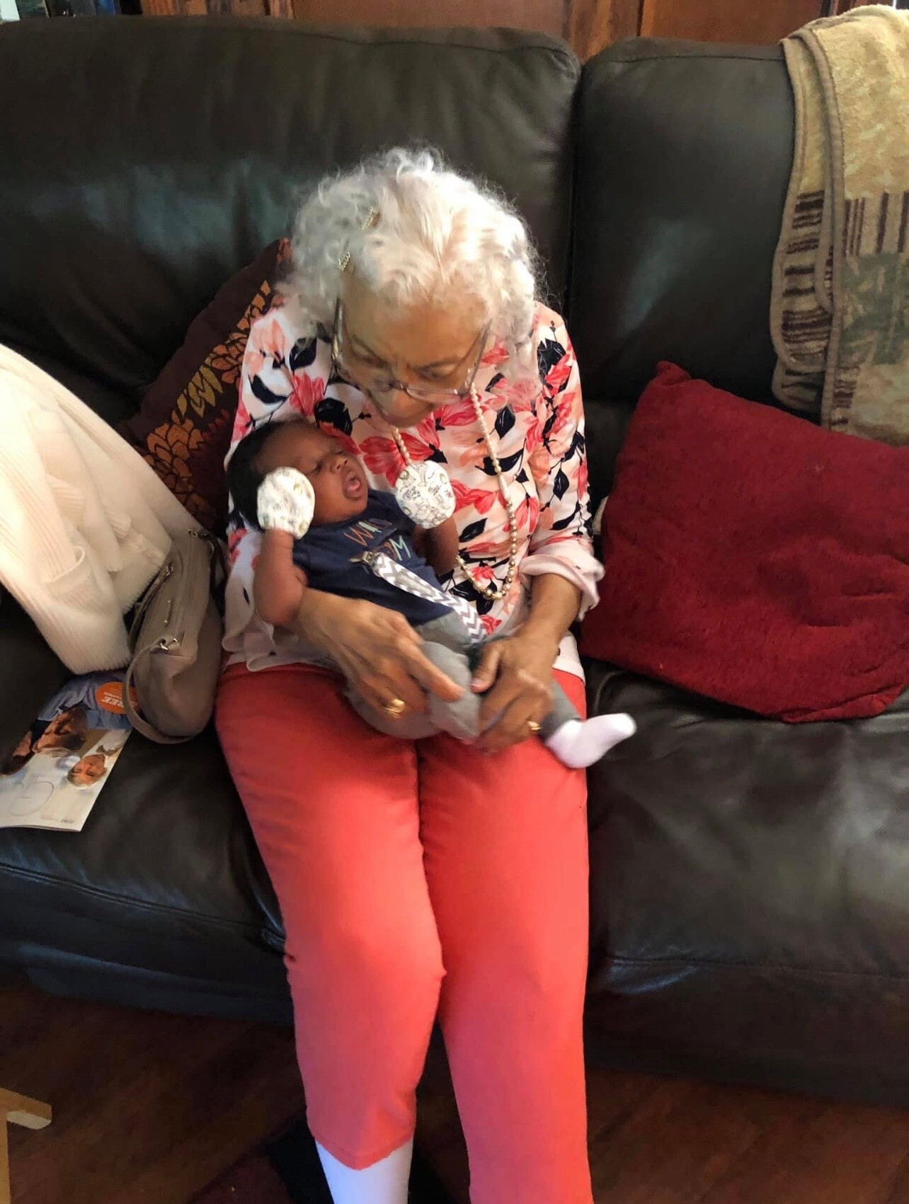 Grandmother with white hair sitting on a black leather couch, cradling a baby in her lap.