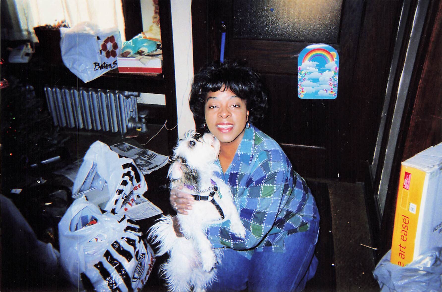 Smiling woman in a blue plaid shirt holding a small white dog indoors among gift bags and wrapping paper.