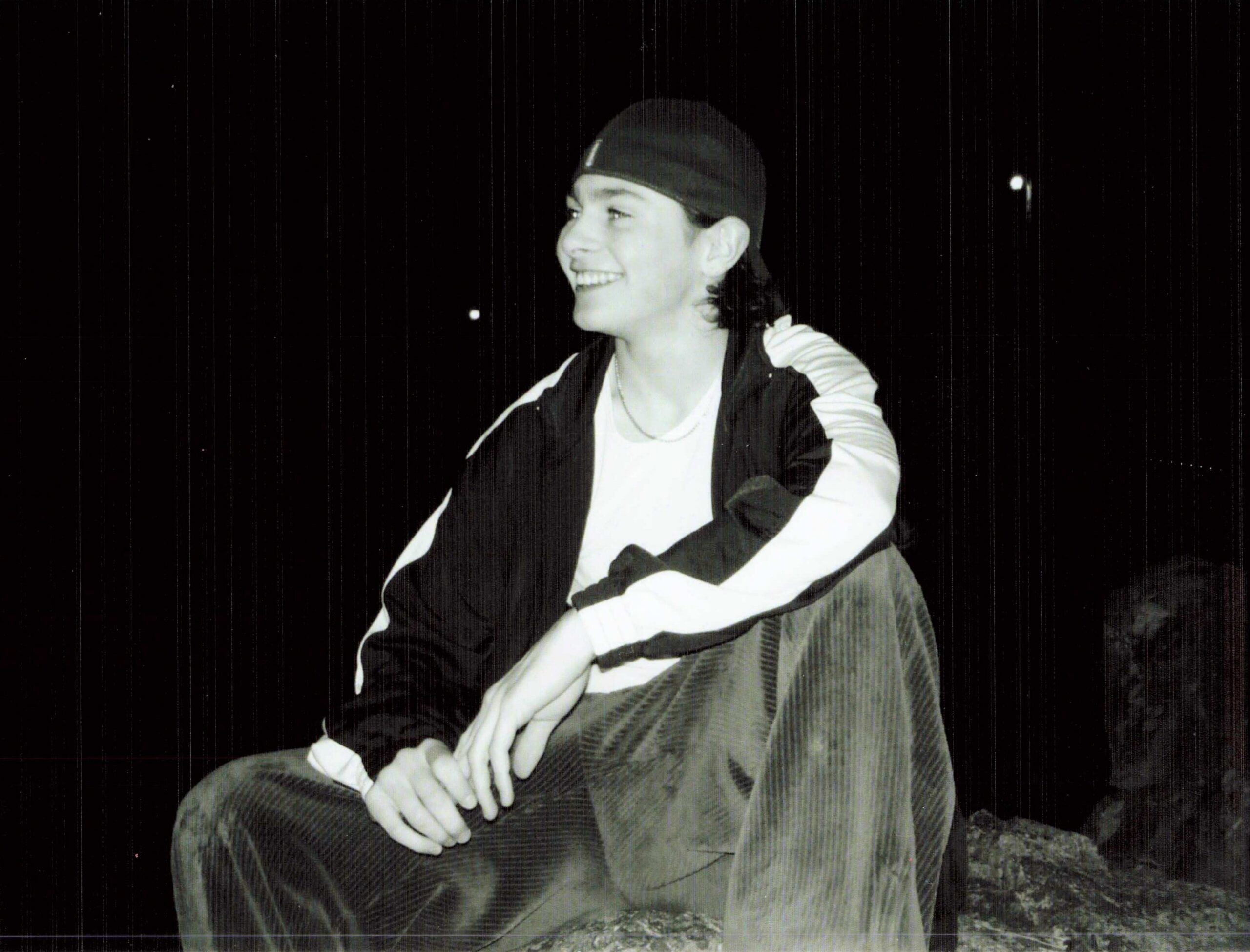 Young person wearing a backward black cap, sitting on a rock at night, smiling.