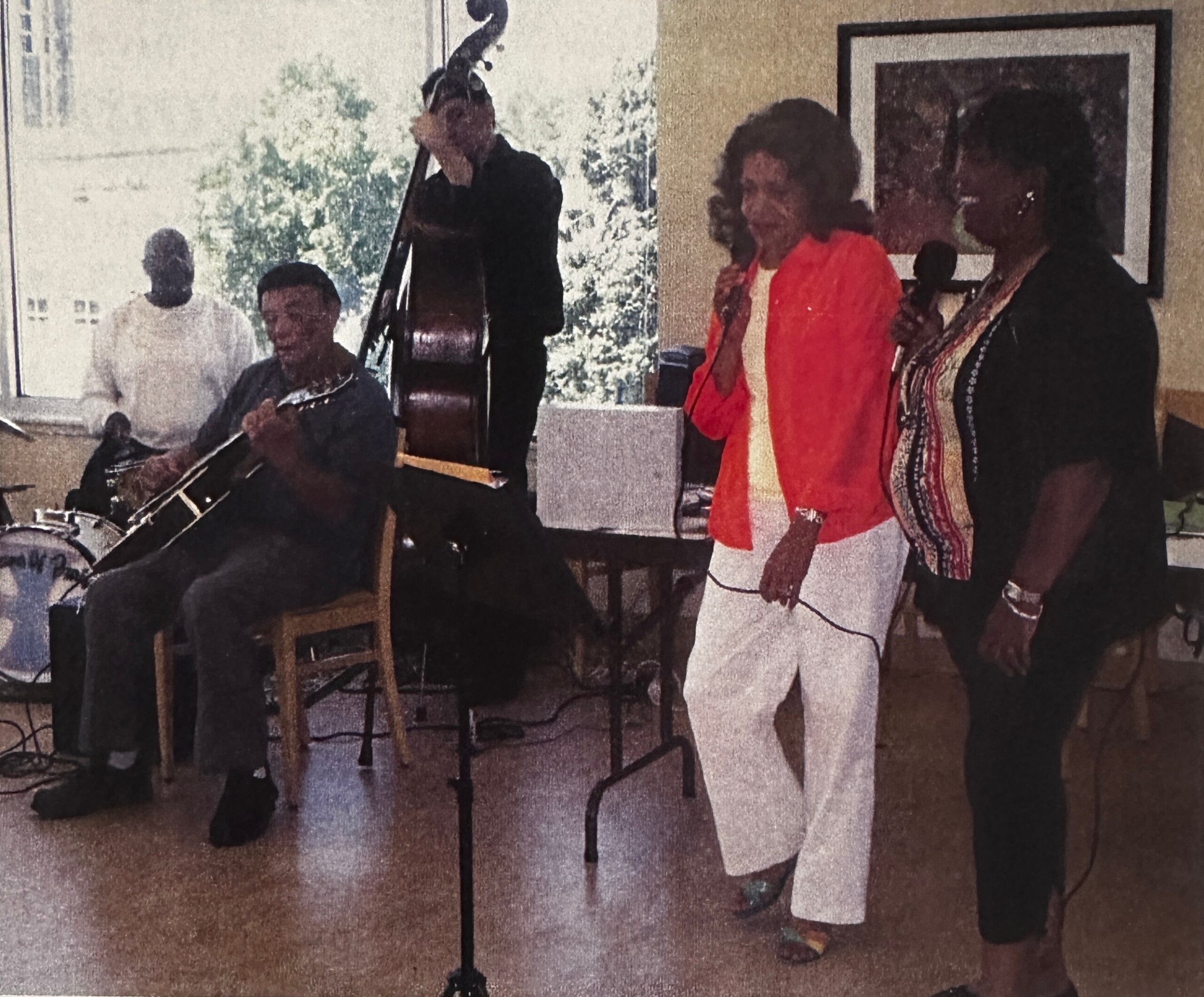 A small band performs indoors with two guitarists and a vocalist, others in the background watching and singing along.