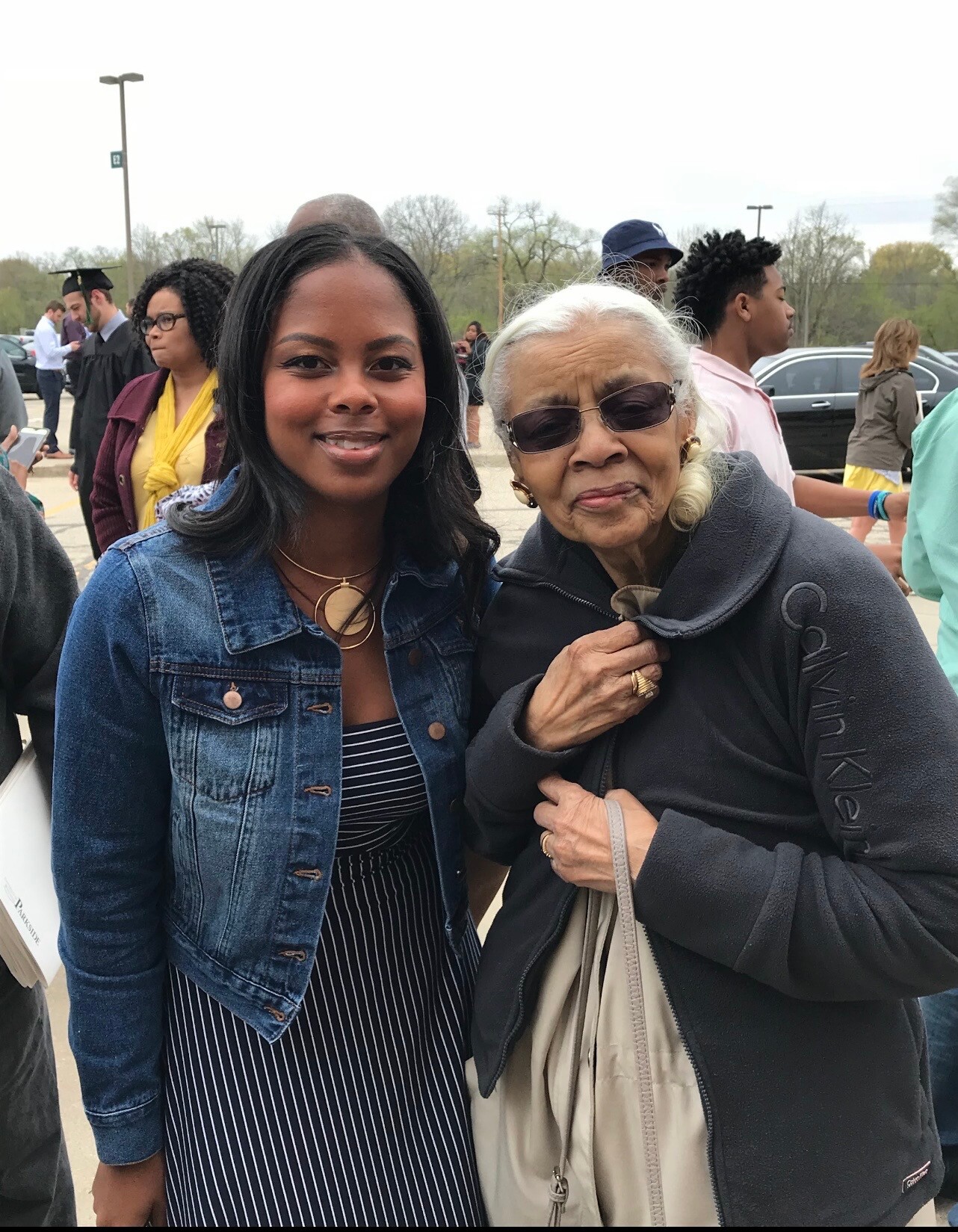 Smiling young woman in a denim jacket posing with an older woman wearing sunglasses at a graduation event.