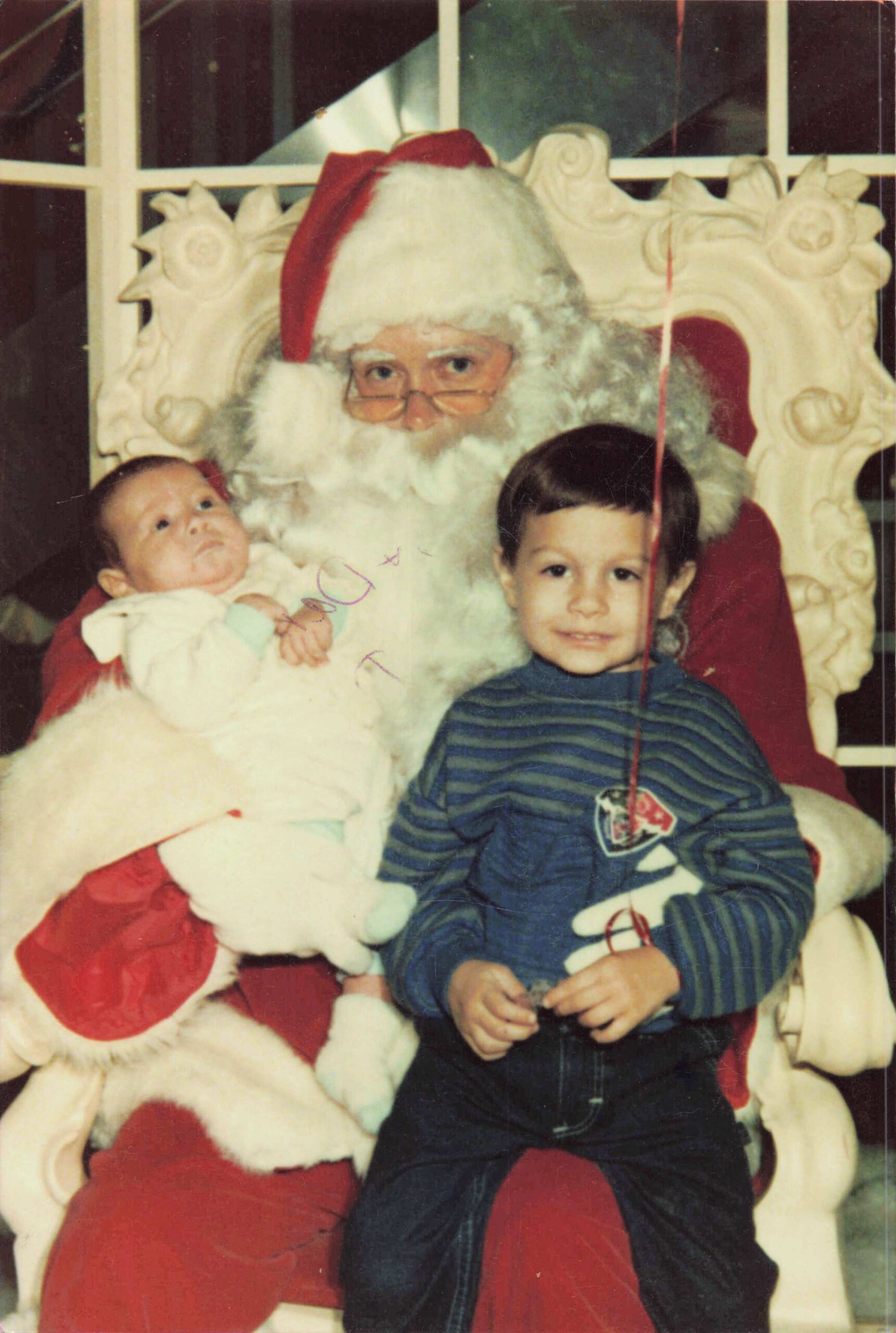 Santa Claus sits in a large ornate chair with a baby in his arms while a young boy sits beside him, posing for a Christmas photo.