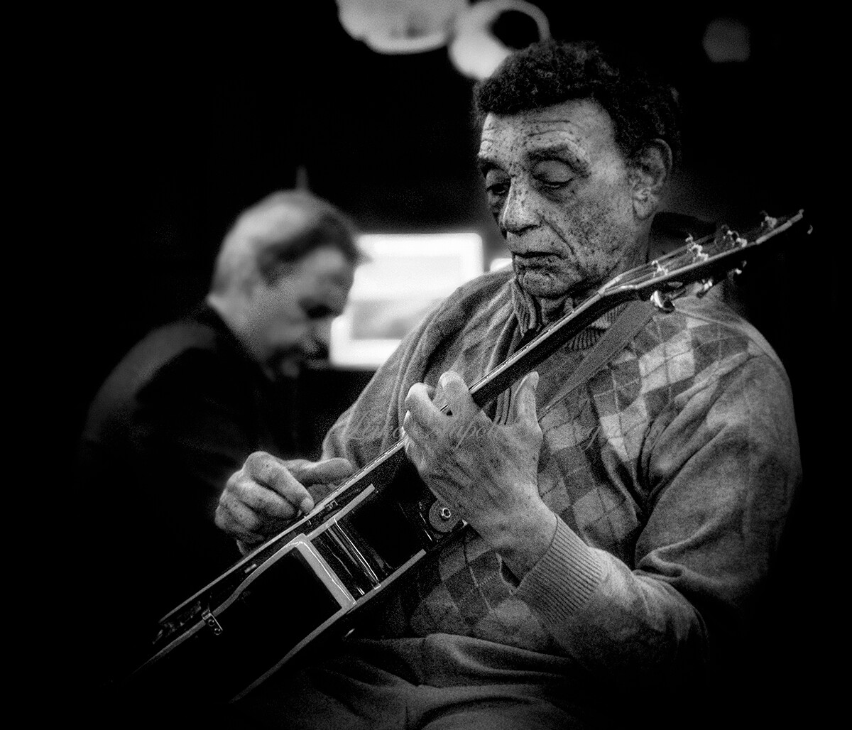 Man playing an electric guitar, eyes closed, in a dim studio; another musician blurred in the background.