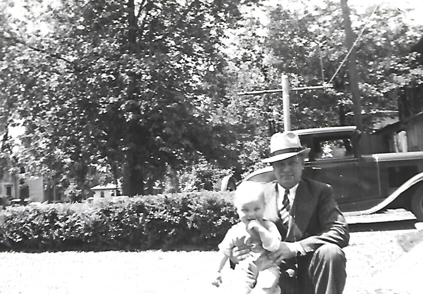 Man in suit and fedora kneels beside a baby outdoors, with a vintage car in the background (black-and-white photo).