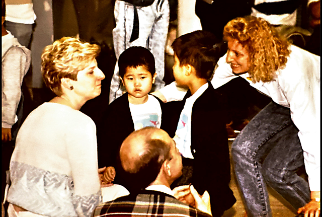 Young boy in a white shirt stands among adults who lean in and talk to him in a crowded room.