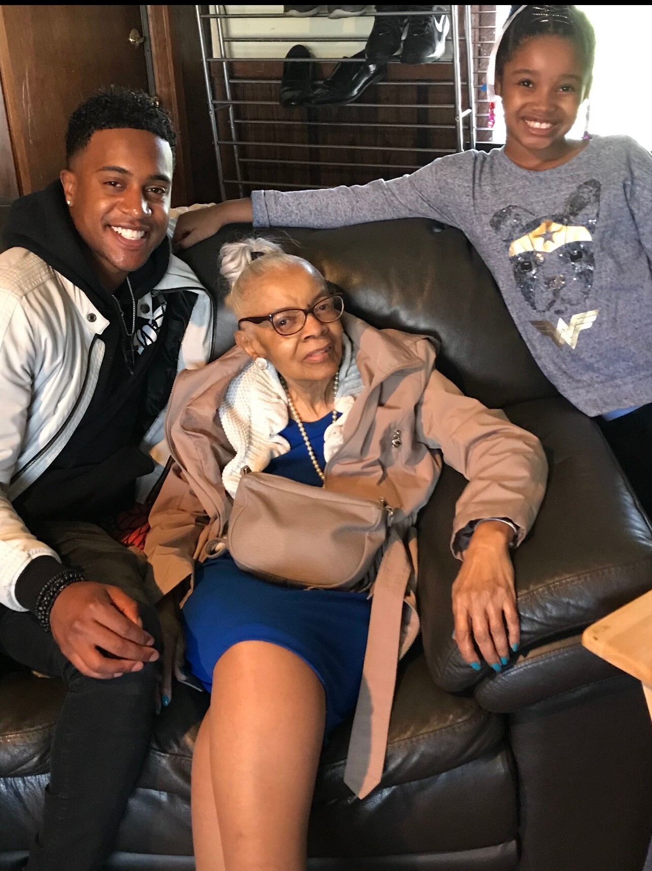 Three people posing on a black leather couch: a smiling young man on the left, an elderly woman with glasses in the middle, and a girl in a gray sweater on the right.