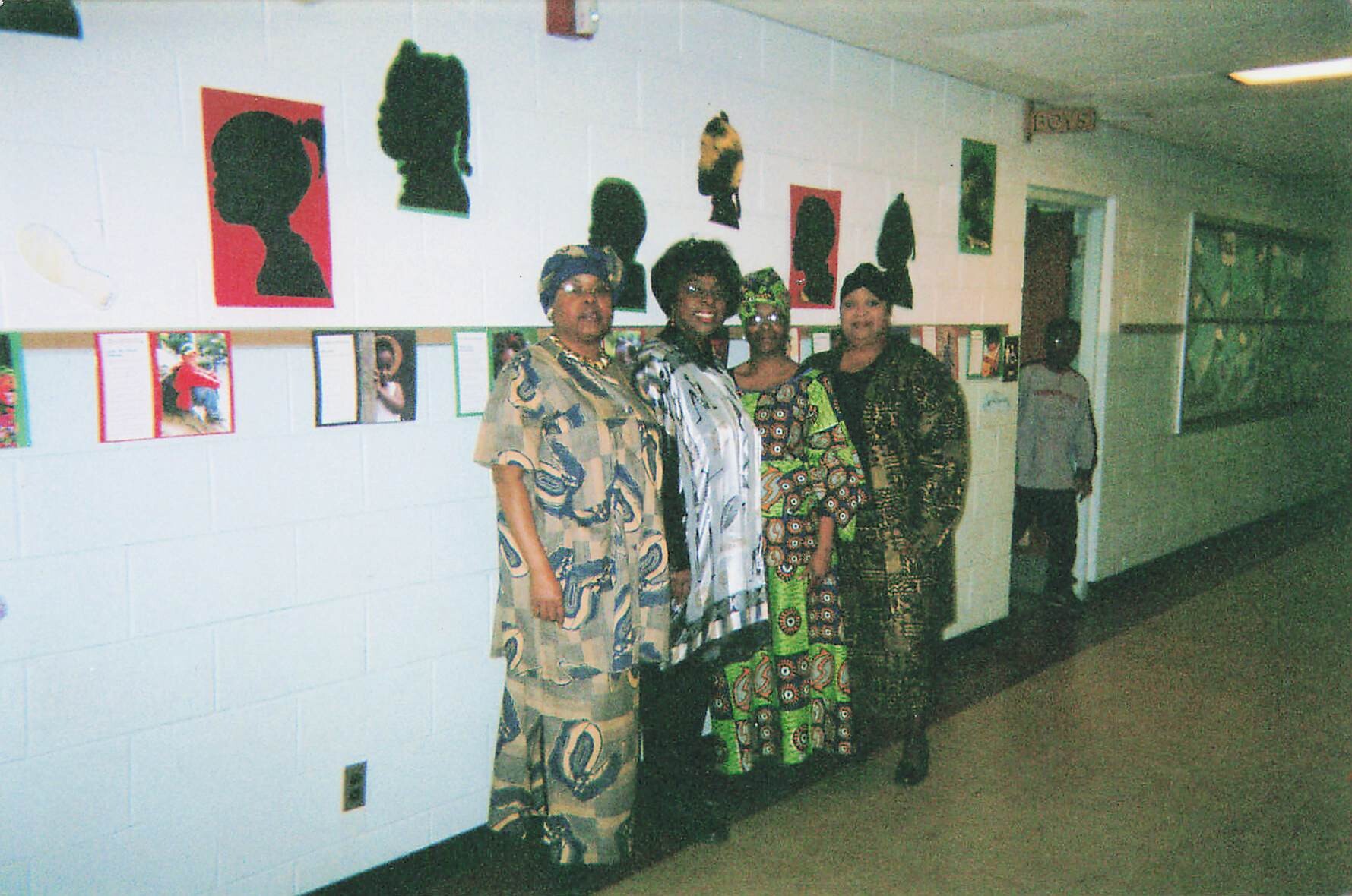 Four women in colorful patterned dresses posing together in a school hallway with artwork silhouettes on the wall behind them