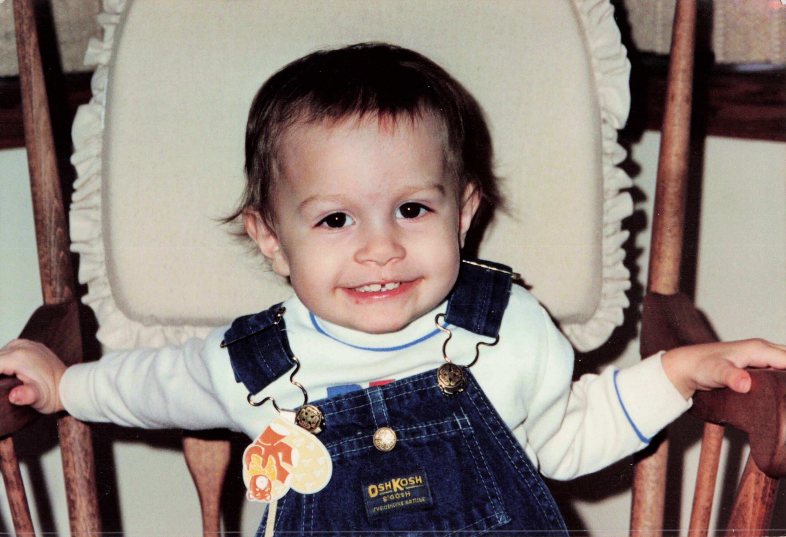 Smiling toddler in blue denim overalls and light shirt sits in a wooden chair with arms raised.