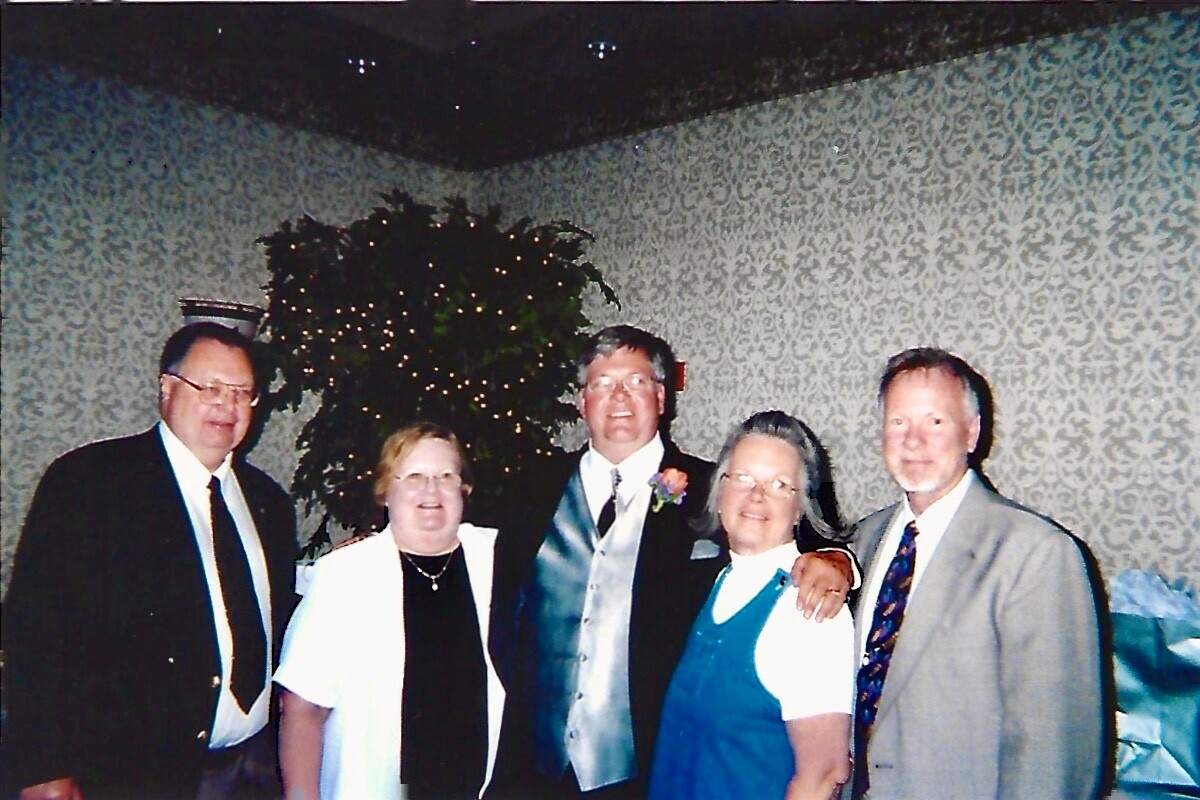 Five adults in formal attire posing together at an indoor event, with a decorative patterned wall in the background and a lit plant behind them.