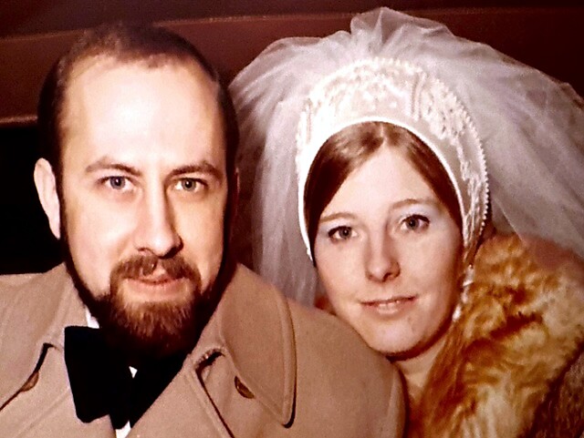Bride and groom posing closely, the bride in a lace veil and fur-trimmed coat beside the groom in a tan coat and black bow tie.