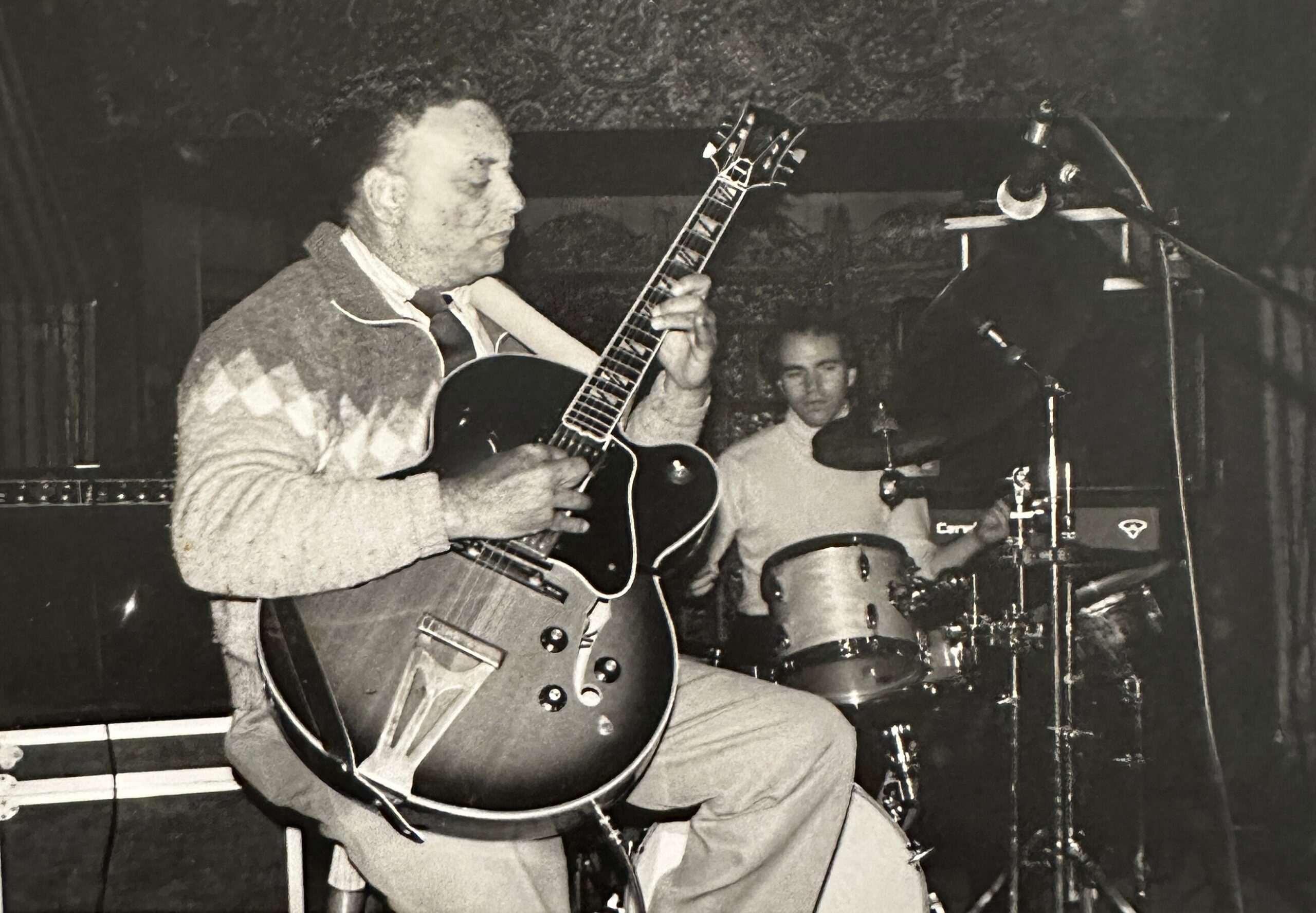 Black-and-white photo of a guitarist in a sweater playing an electric guitar on a dim stage, with a drummer in the background.
