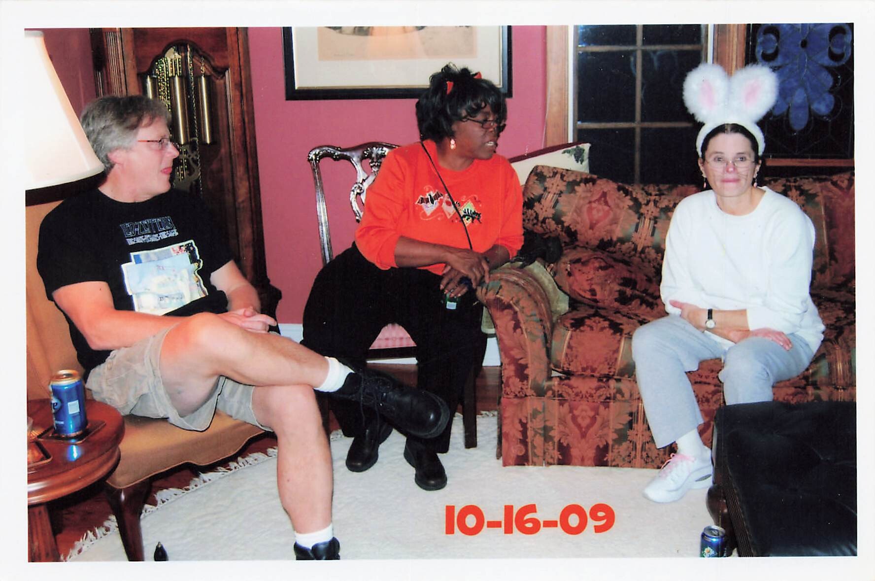 Three people sit in a living room chatting: left man in black T-shirt, center woman in orange, right woman wearing fluffy bunny ears; date 10-16-09 on the floor visible below.