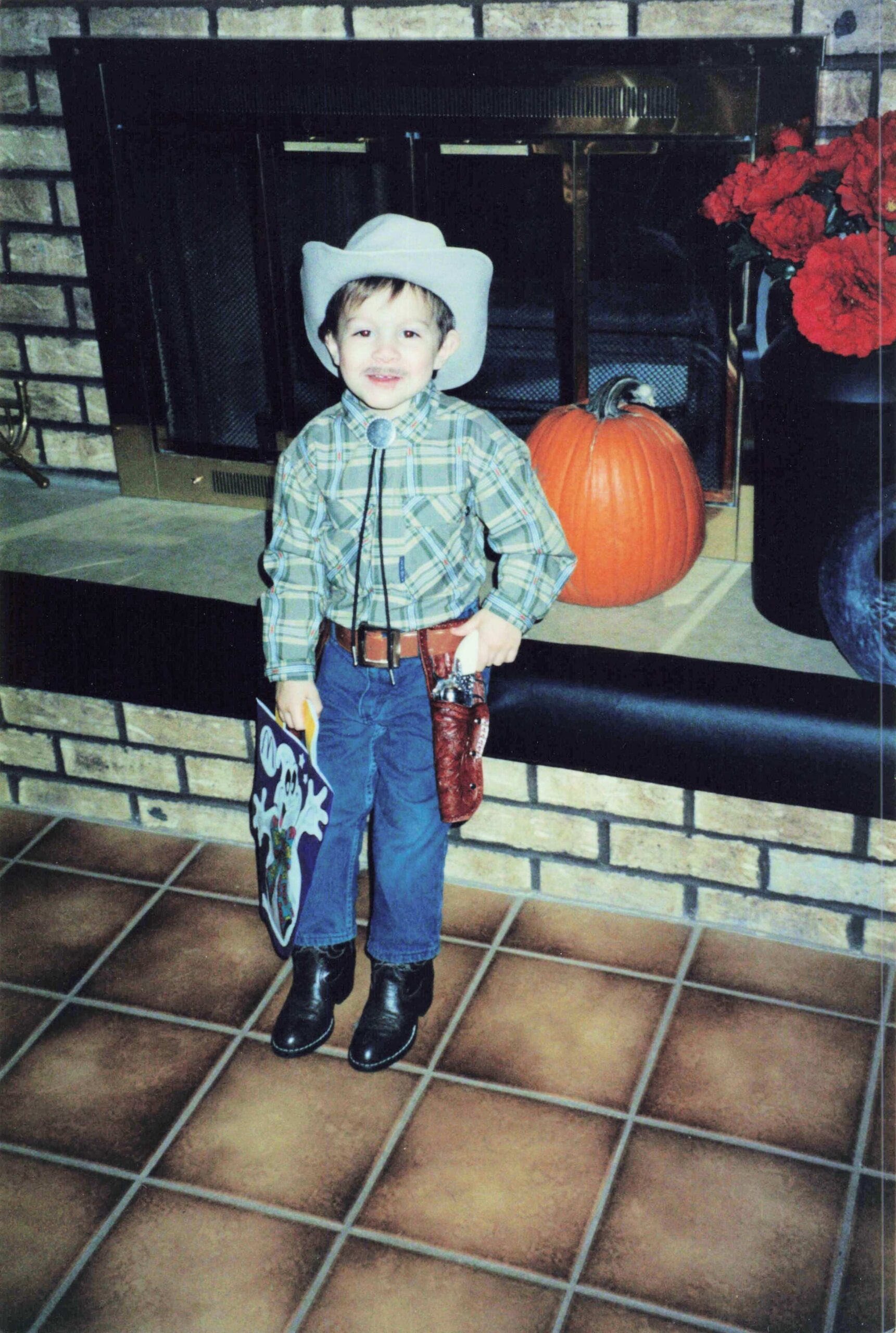 Boy dressed as a cowboy in a plaid shirt and hat standing in front of a fireplace with a pumpkin beside him.