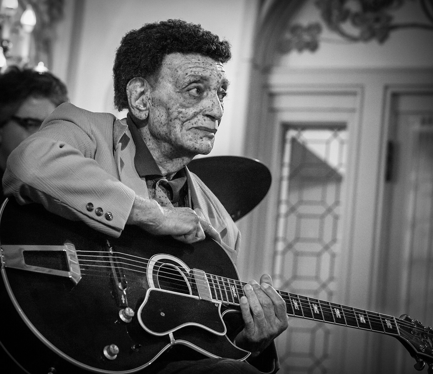 Senior man in a blazer playing an acoustic guitar, looking thoughtful in a home setting (black and white photo).