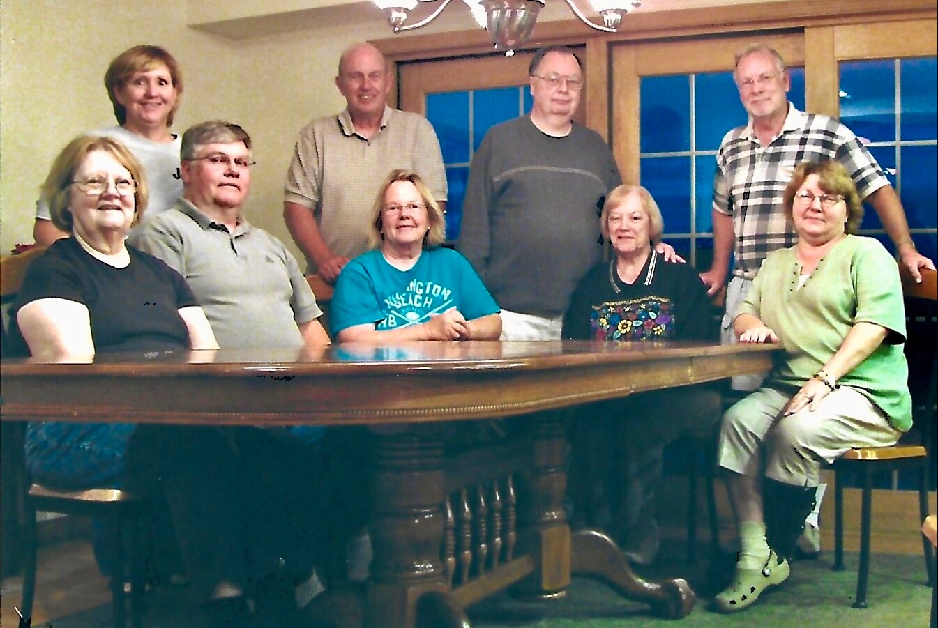 Nine adults gathered around a large wooden table, posing for a casual group photo in a dining room.