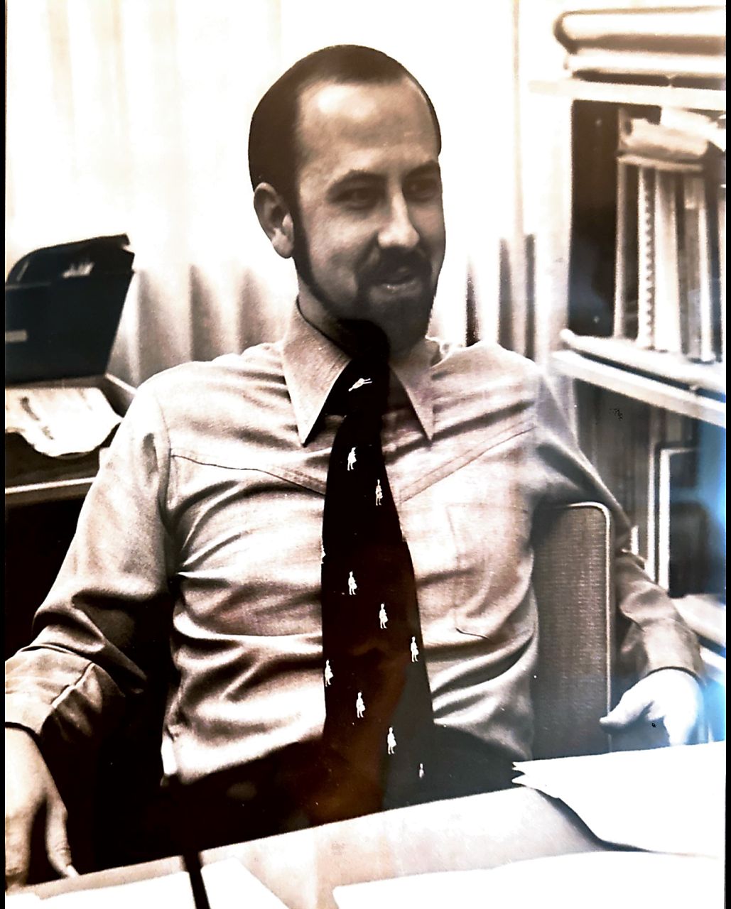 Man seated at a desk in an office, wearing a light shirt and a tie with small white figures.