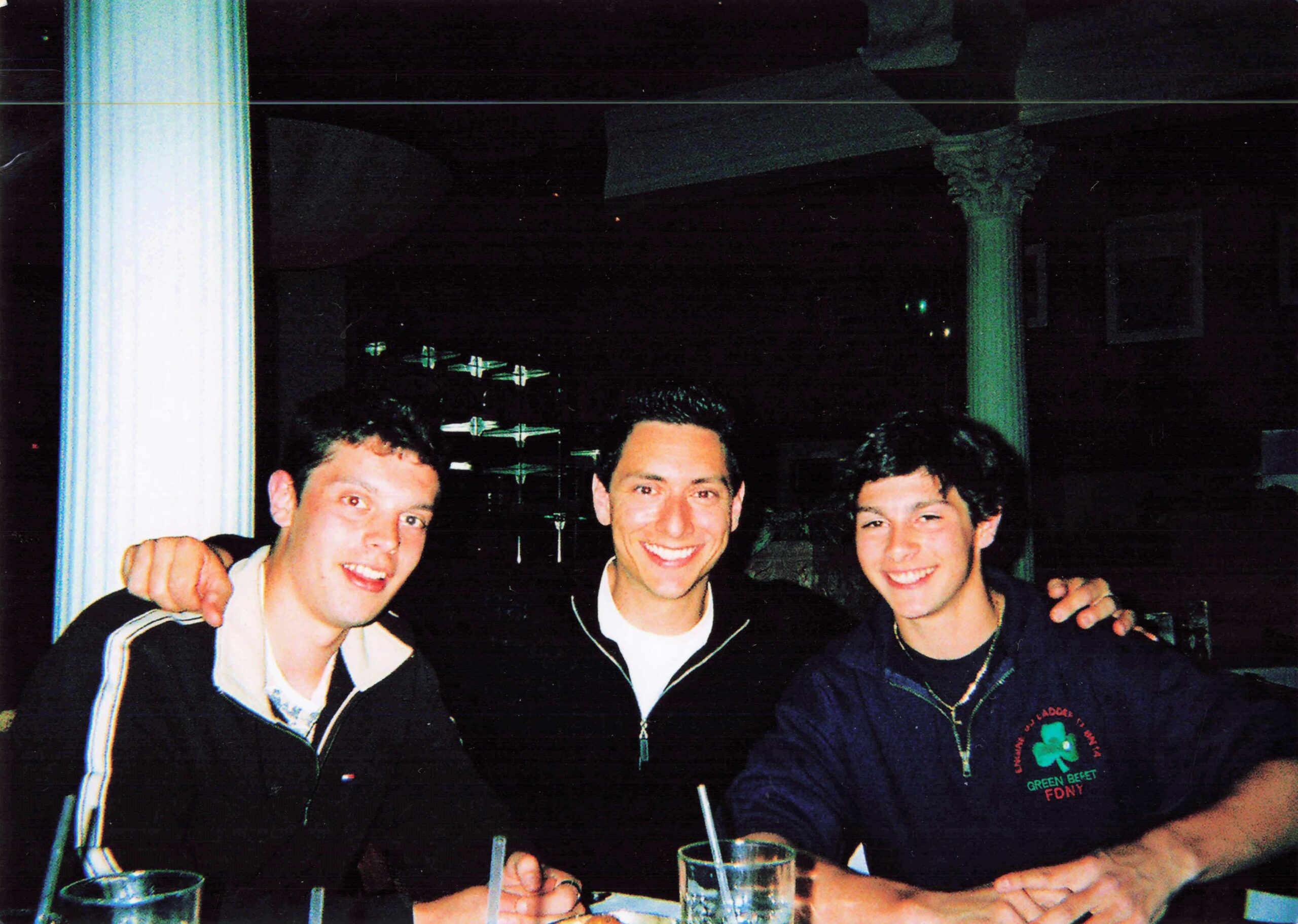 Four men pose together for a casual group photo at a nighttime gathering, with drinks on the table in front of them, smiling at the camera.