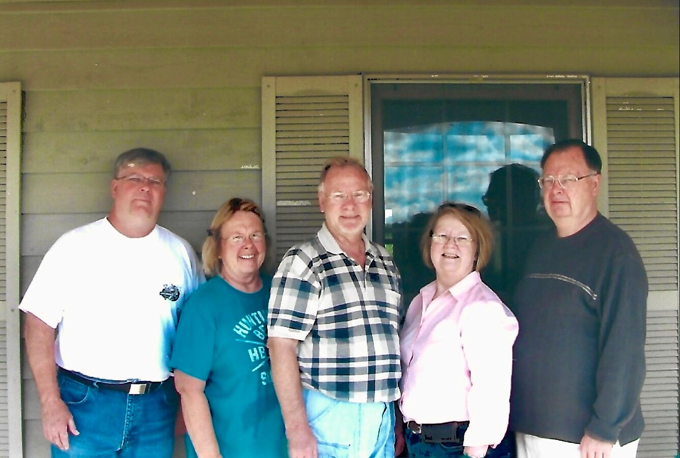 Five adults standing together outside a house, posing in front of a window with shutters and smiling for the camera.