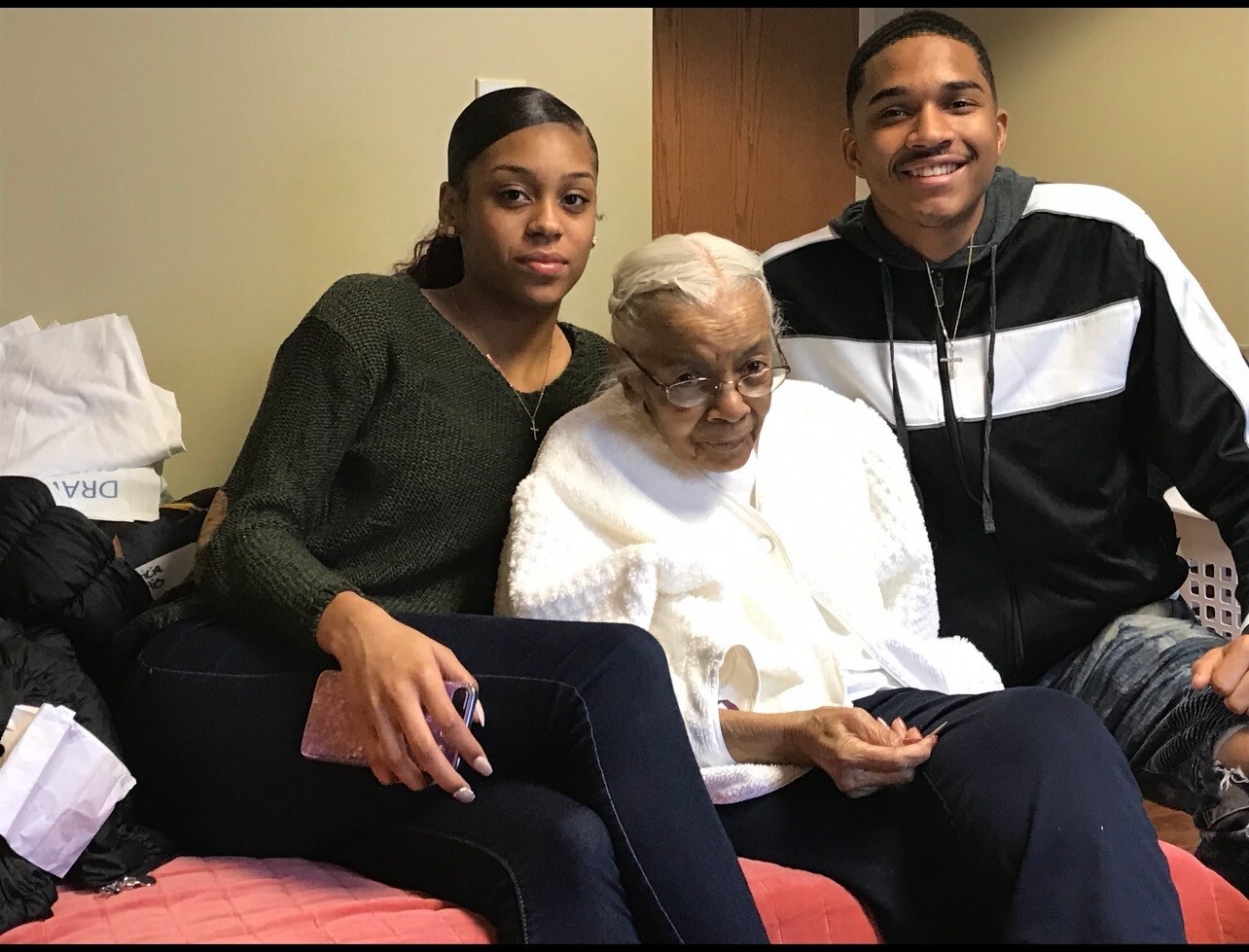 Three generations sit together on a couch: a young woman left, an elderly woman in a white coat center, and a smiling young man right.