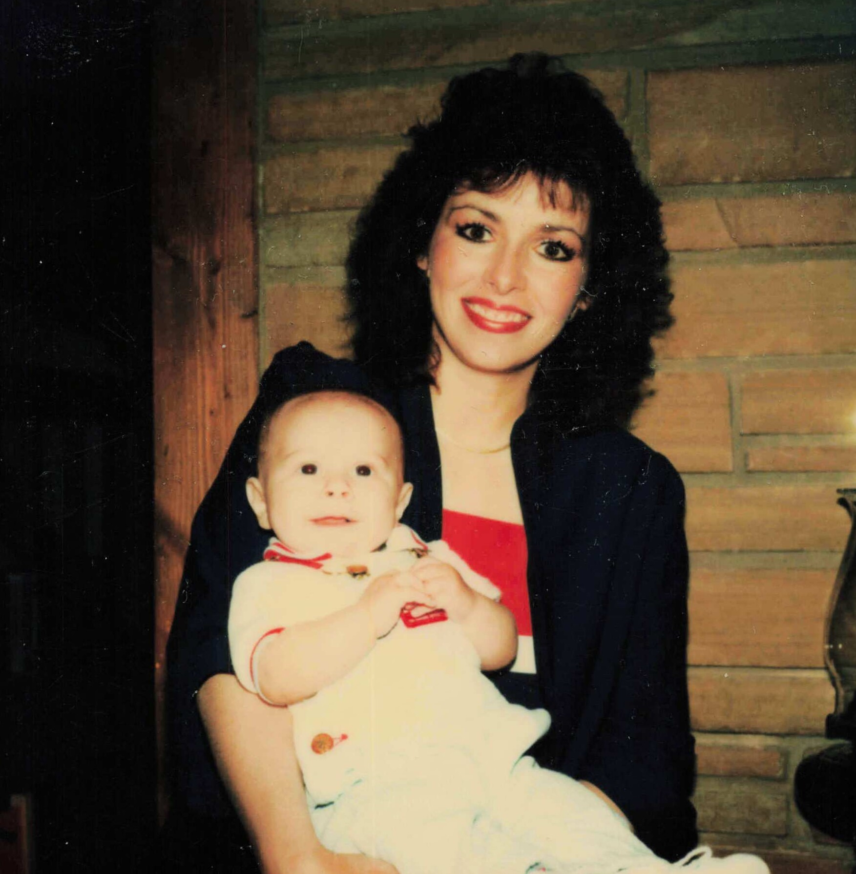 Woman with dark hair smiling while holding a baby in a red and white outfit, against a brick wall background.