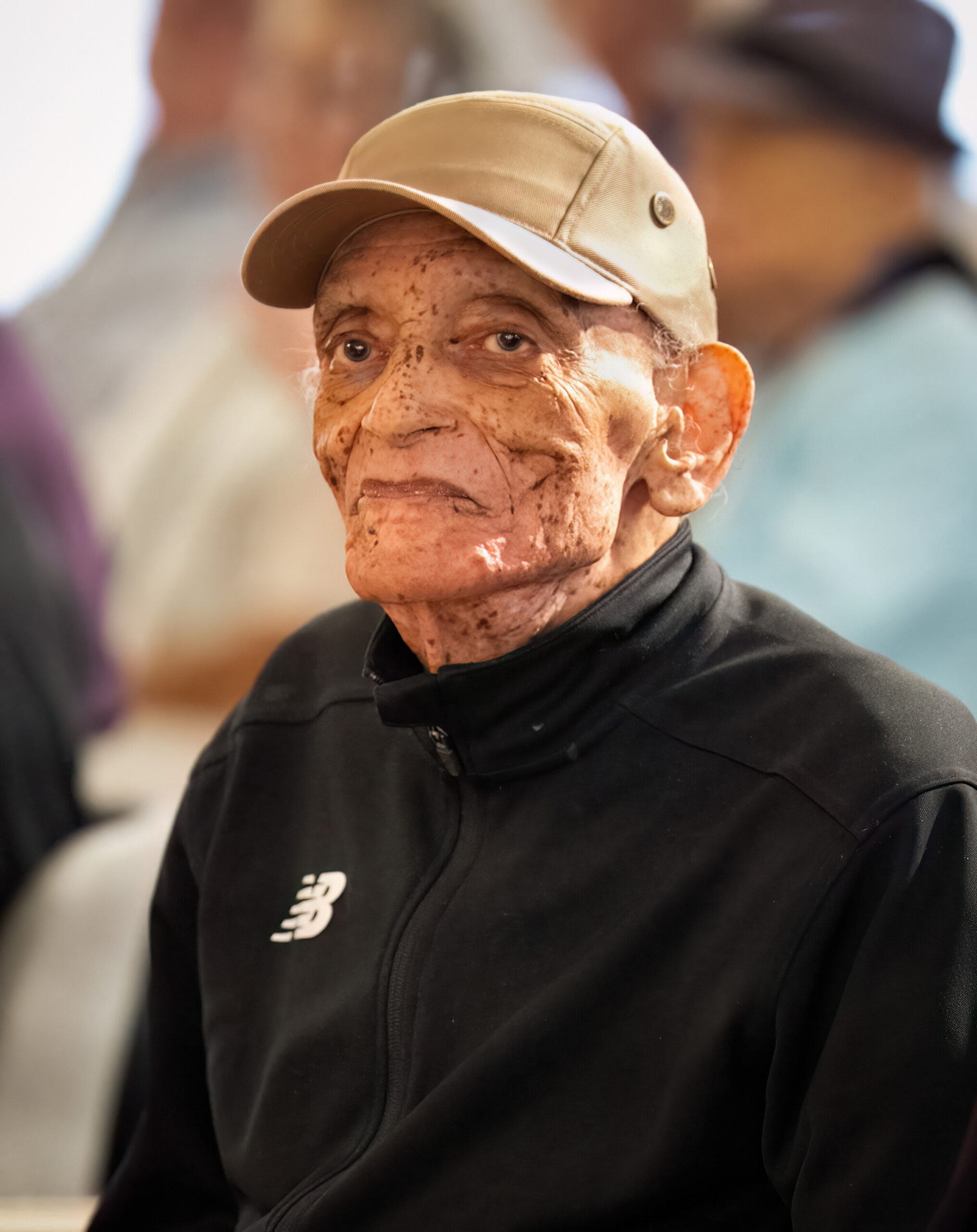 Elderly man with a beige cap and black zip-up jacket, close-up portrait.