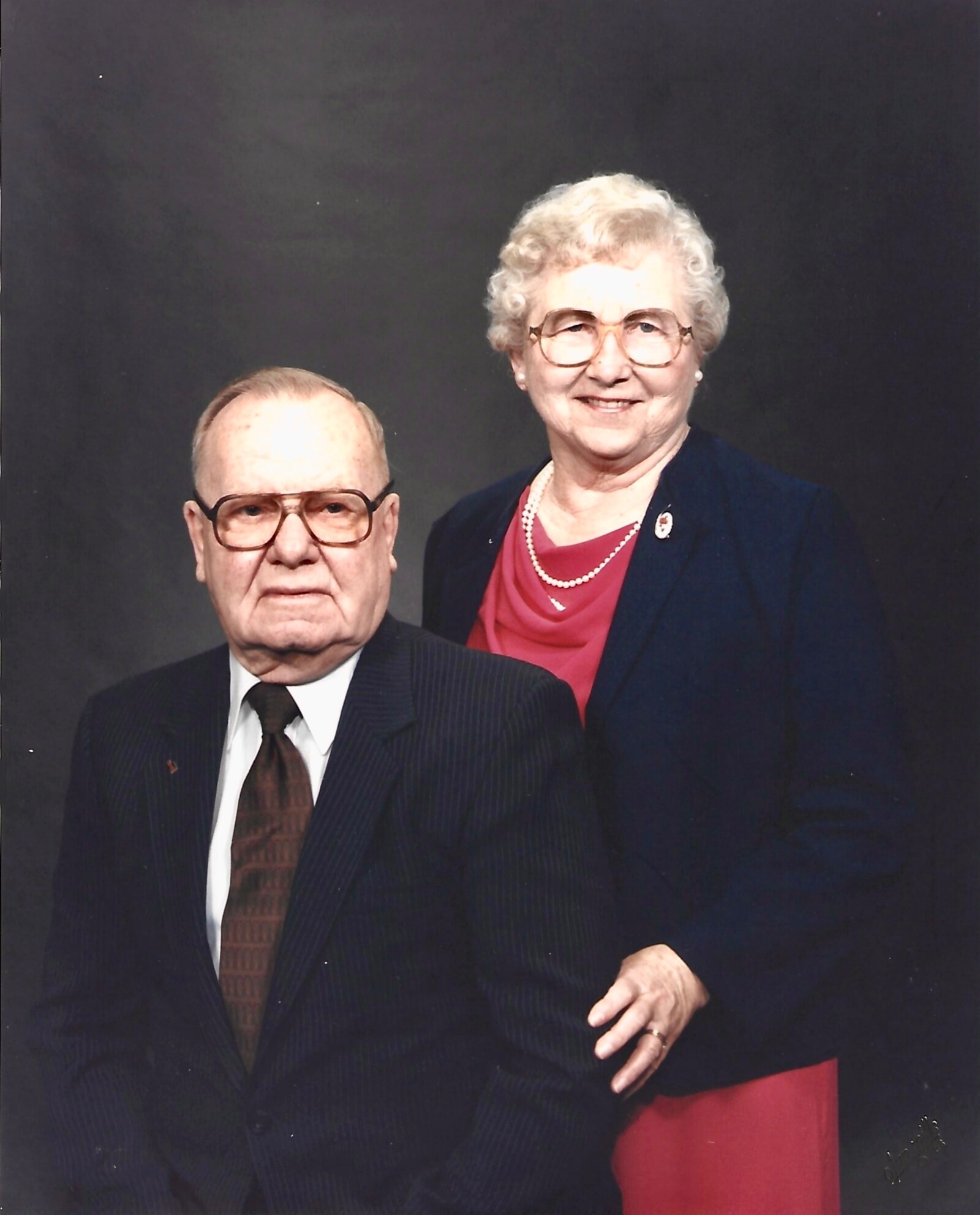 Elderly couple posing for a formal portrait: man seated in a pinstriped suit and brown tie, woman standing behind him in a navy blazer with a red blouse and pearls, both smiling.