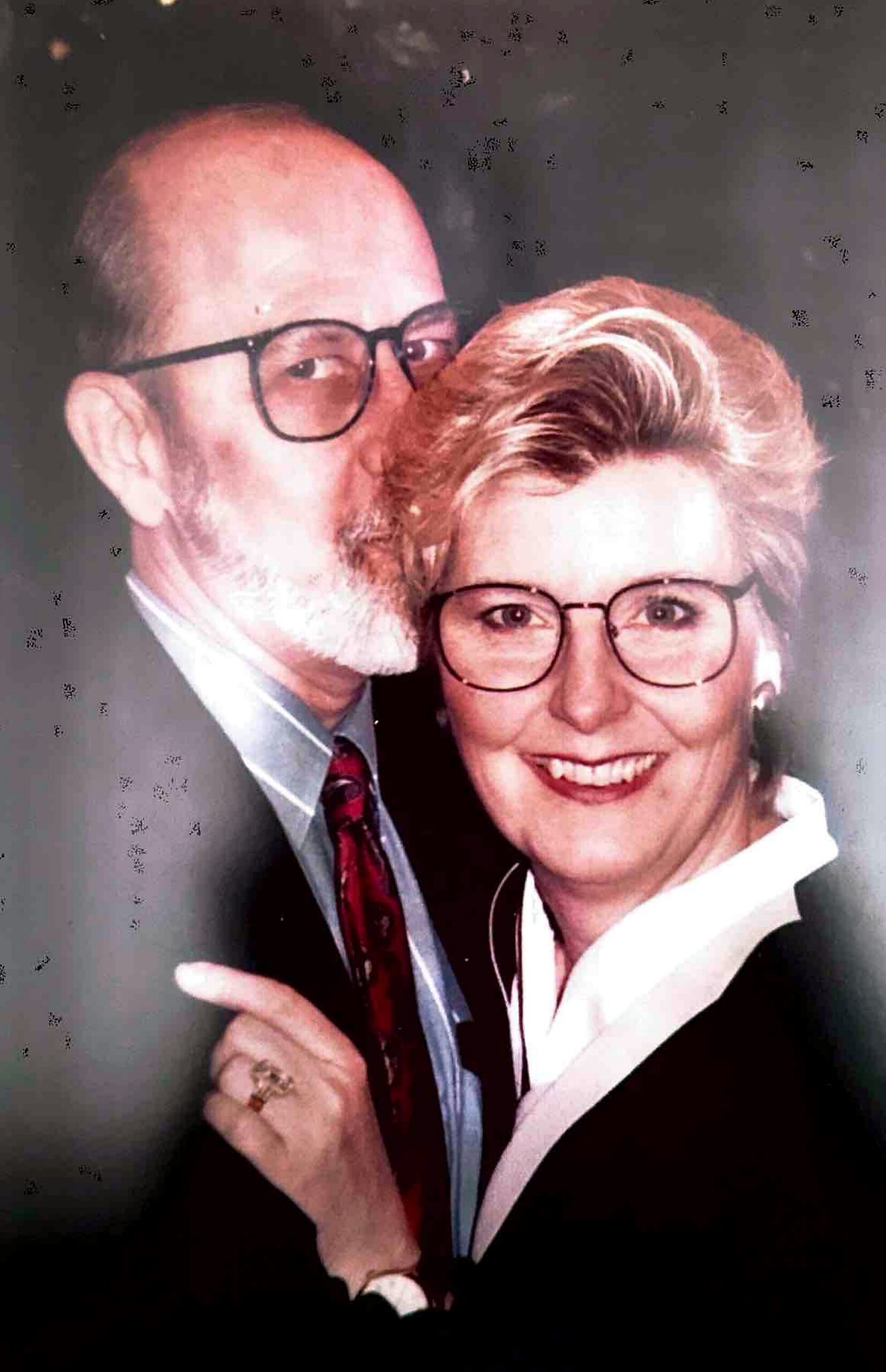 Older couple pose for a photo: man kissing the woman on the cheek, both wearing glasses and smiling beneath formal attire.