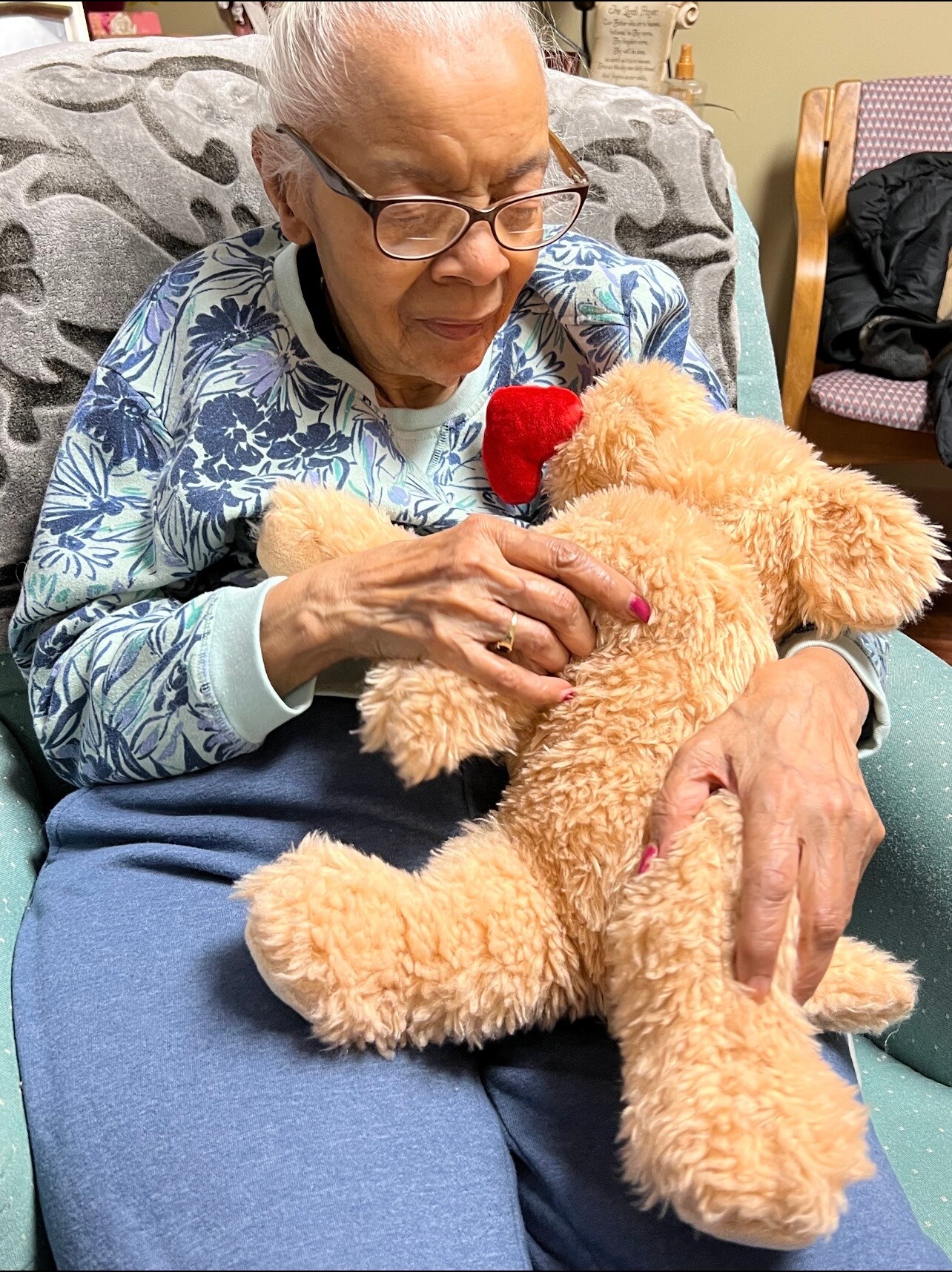 Senior woman with glasses sits on a sofa, cuddling a fluffy teddy bear with a red heart close to her chest.