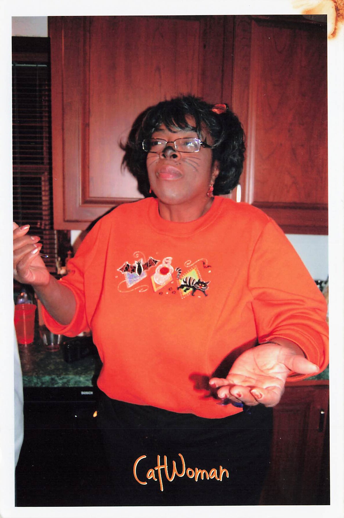 Woman wearing an orange shirt with a CatWoman graphic, glass-wearing and face paint whiskers, arms raised in a kitchen with wooden cabinets behind her.