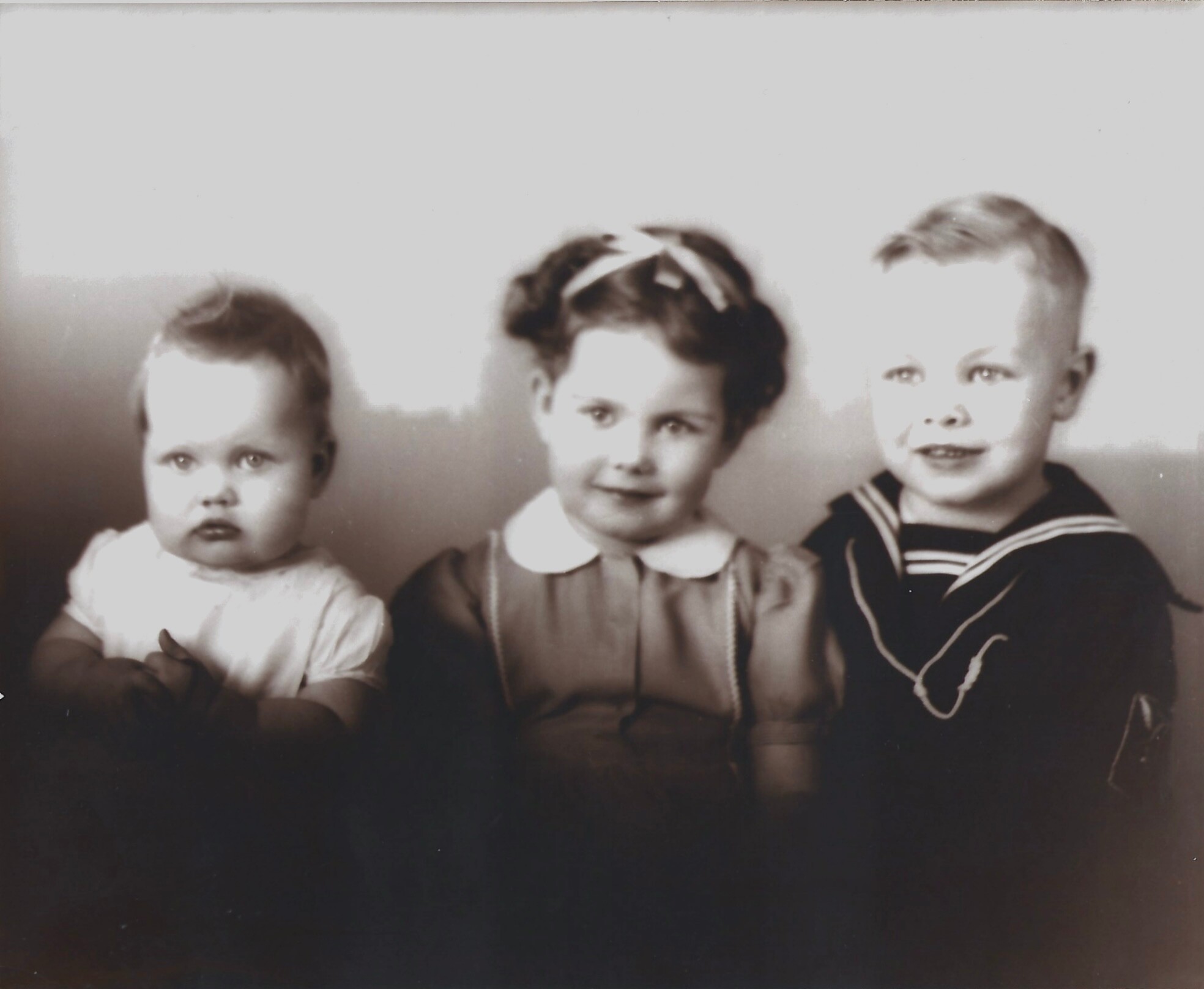 Three young children posing for a vintage studio portrait: toddler on the left, a girl with a bow in the middle, and a boy in a sailor suit on the right.