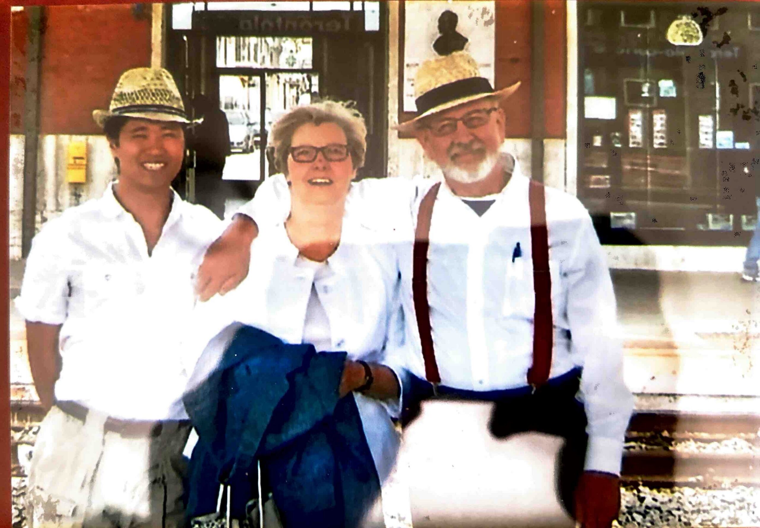 Three adults in white shirts and hats pose arm-in-arm outside a storefront, smiling at the camera.