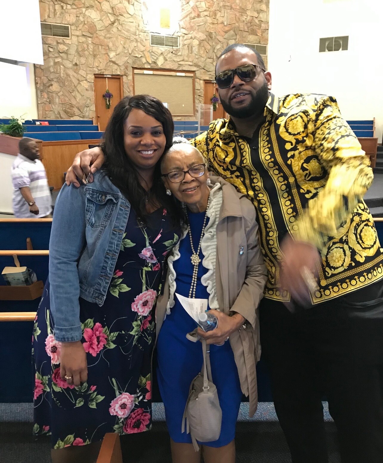 Three people pose together and smile inside a church; a woman in a floral dress and denim jacket stands on the left, an elderly woman in a blue outfit and beige coat in the center, and a man in a gold patterned shirt on the right, with pews and a stone wall in the background.