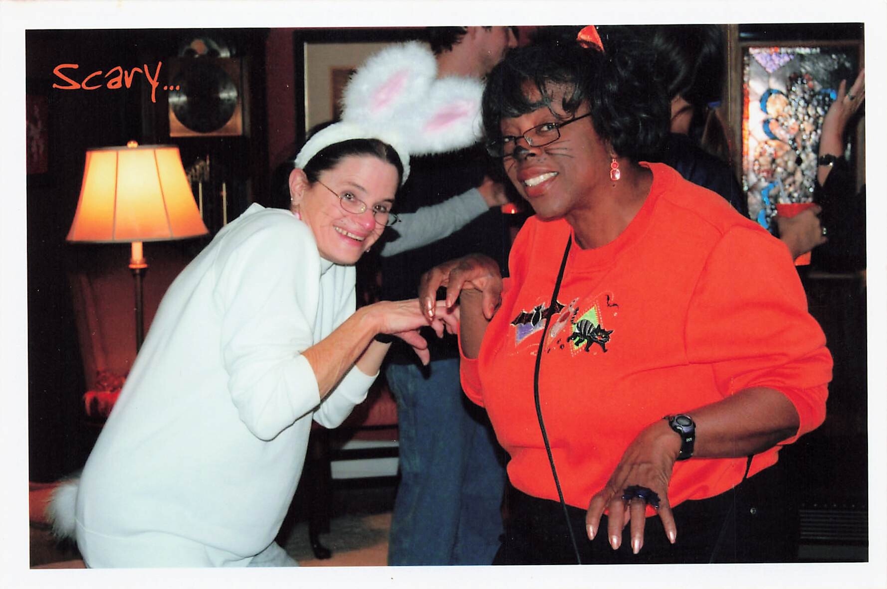 Two people posing playfully at a party: one in white with fluffy bunny ears, the other in an orange top with cat whiskers makeup.