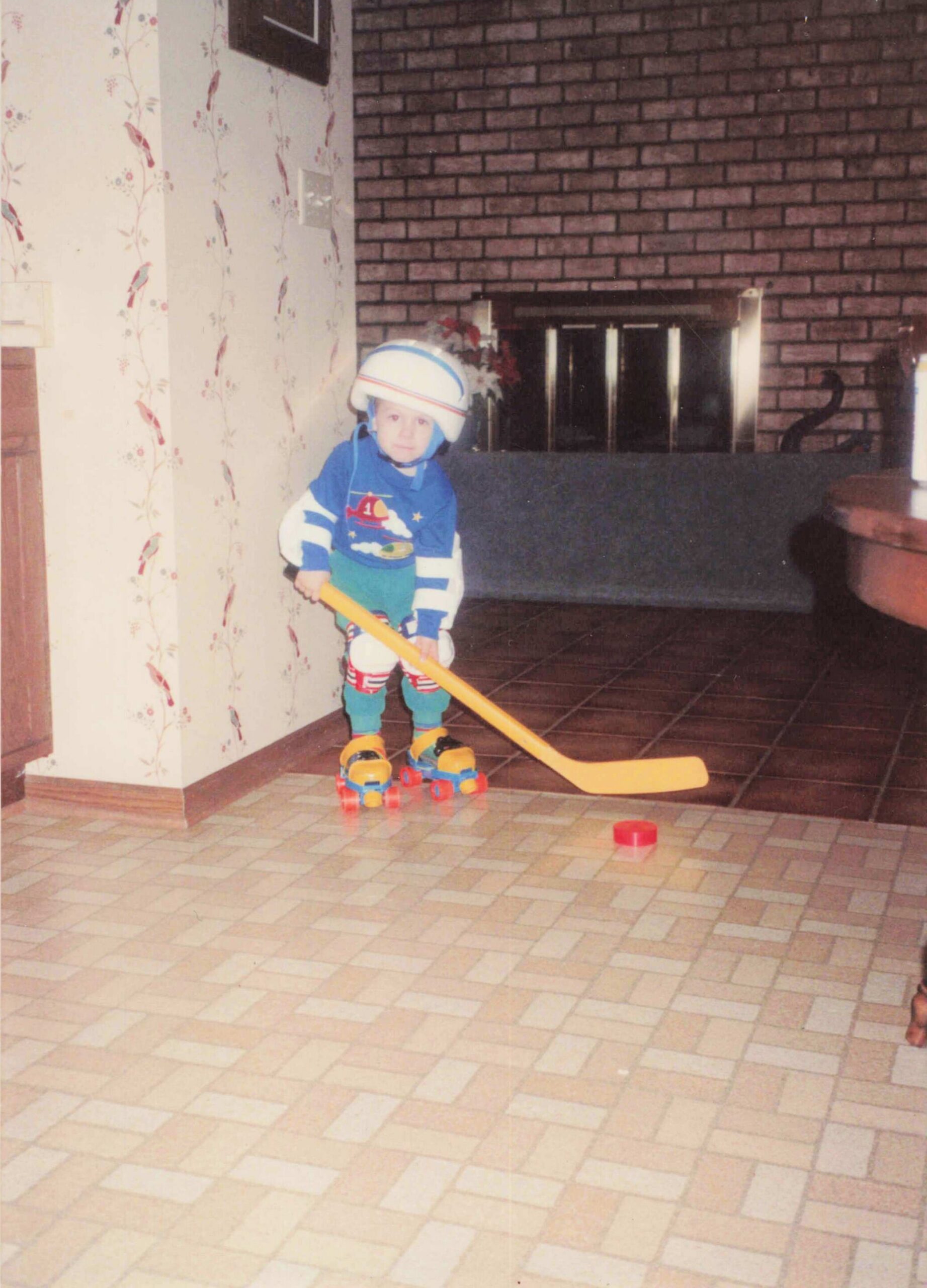 Young child in blue hockey outfit and helmet, holding a yellow hockey stick indoors near a brick fireplace.